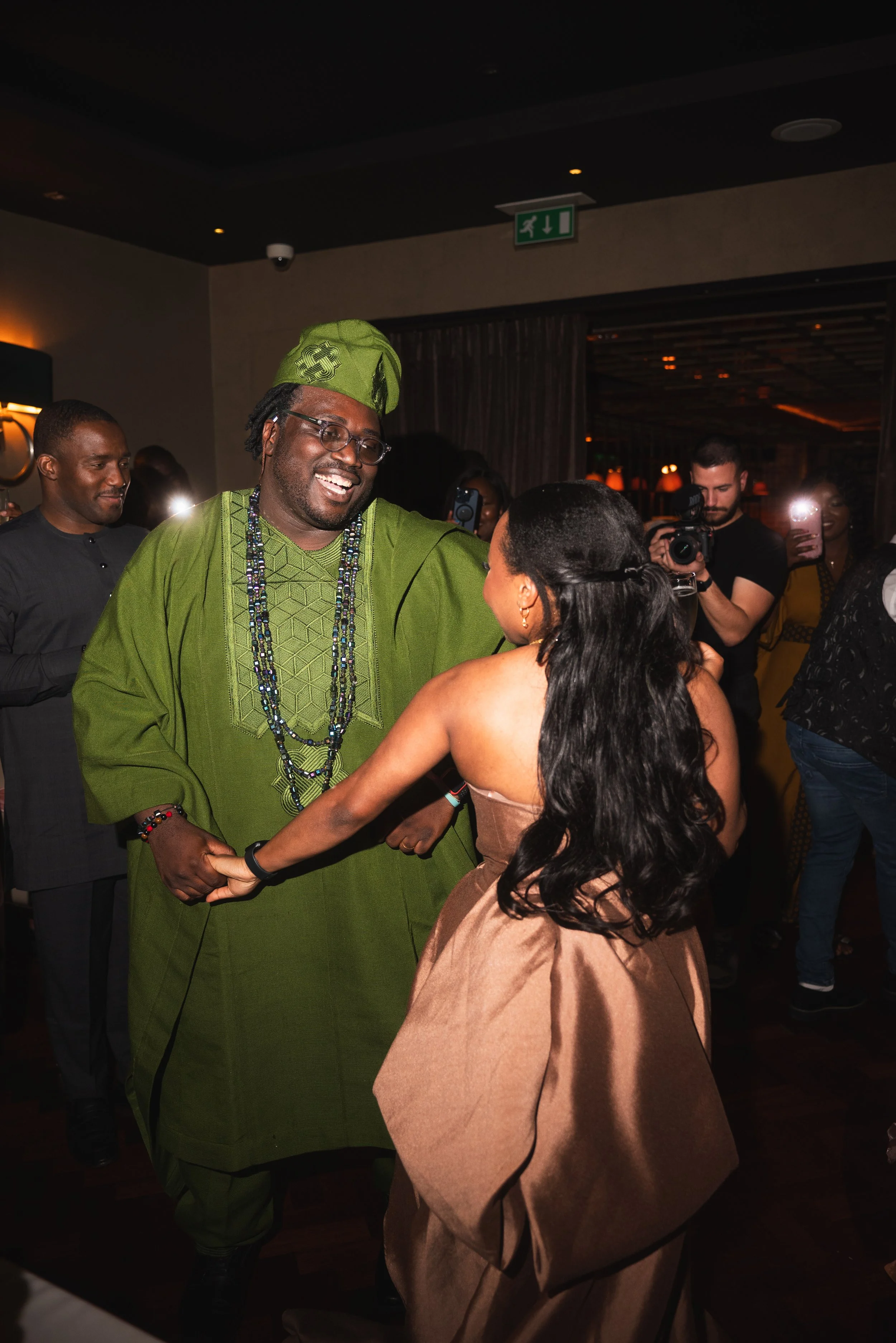 A man wearing a green traditional outfit and headgear dancing with a woman in a brown dress at a celebration or party with people watching and taking photos.
