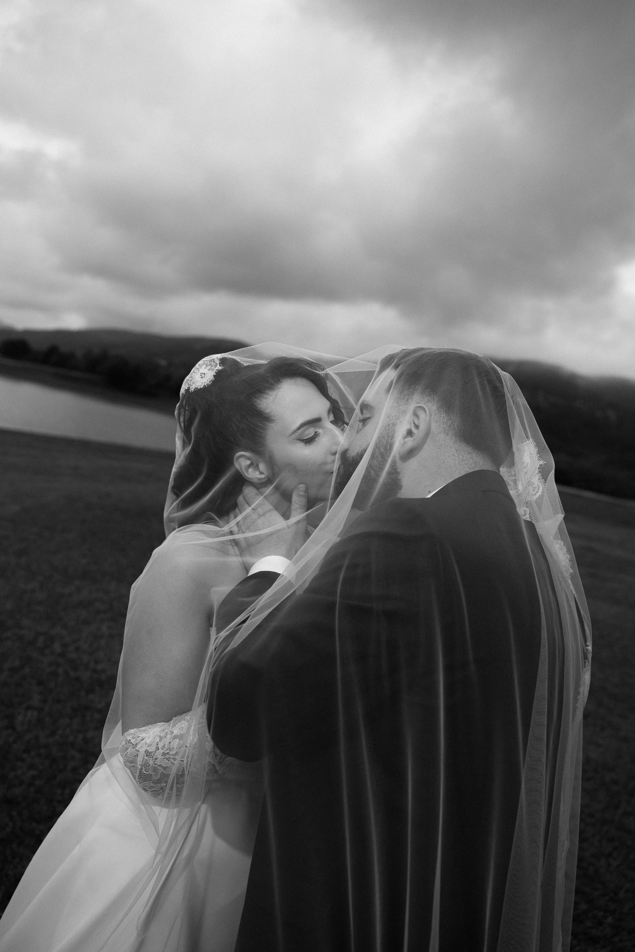 A black and white photo of a bride and groom kissing outdoors under a veil with cloudy sky and hills in the background.