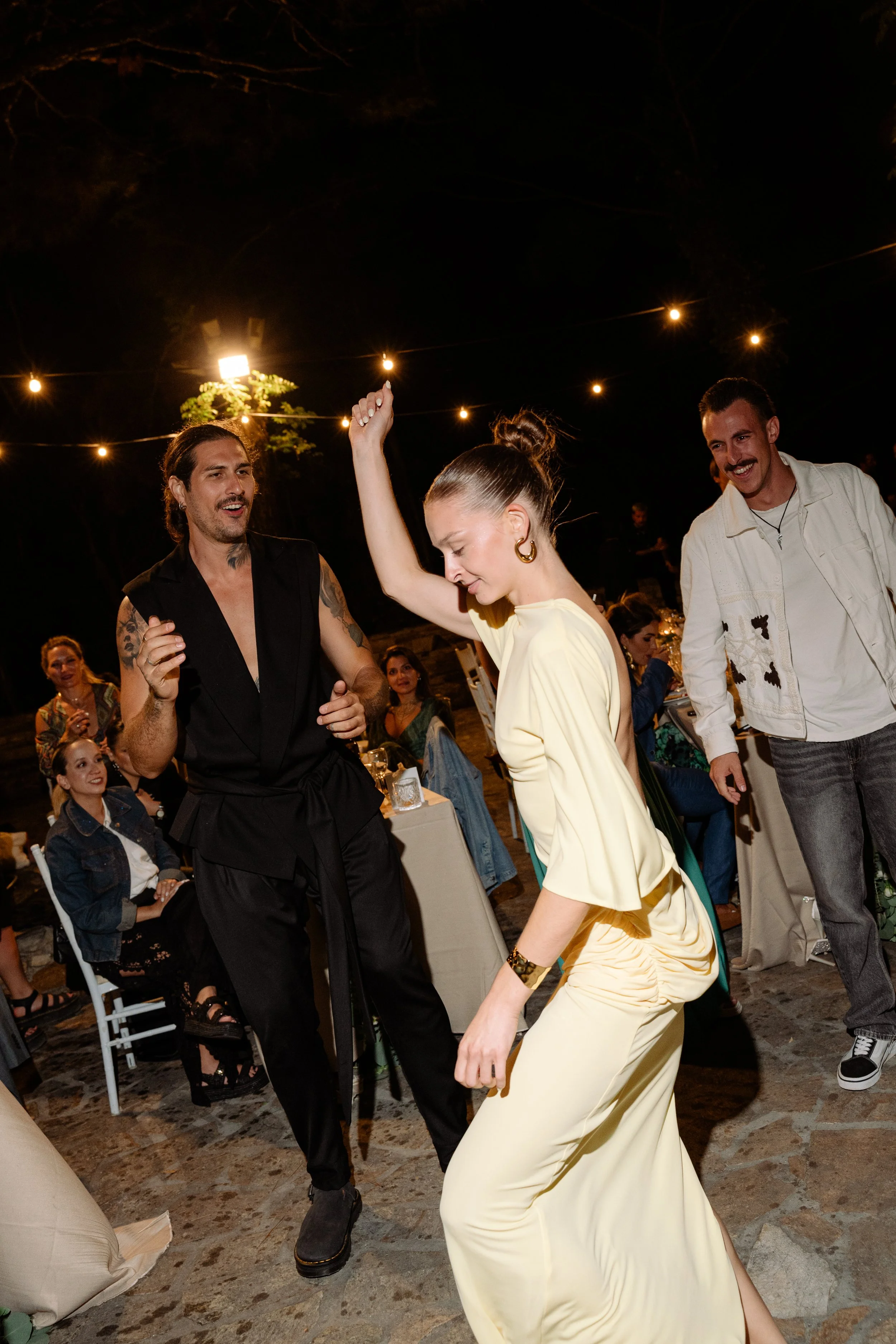 People dancing at an outdoor nighttime celebration under string lights, with some sitting at tables in the background.