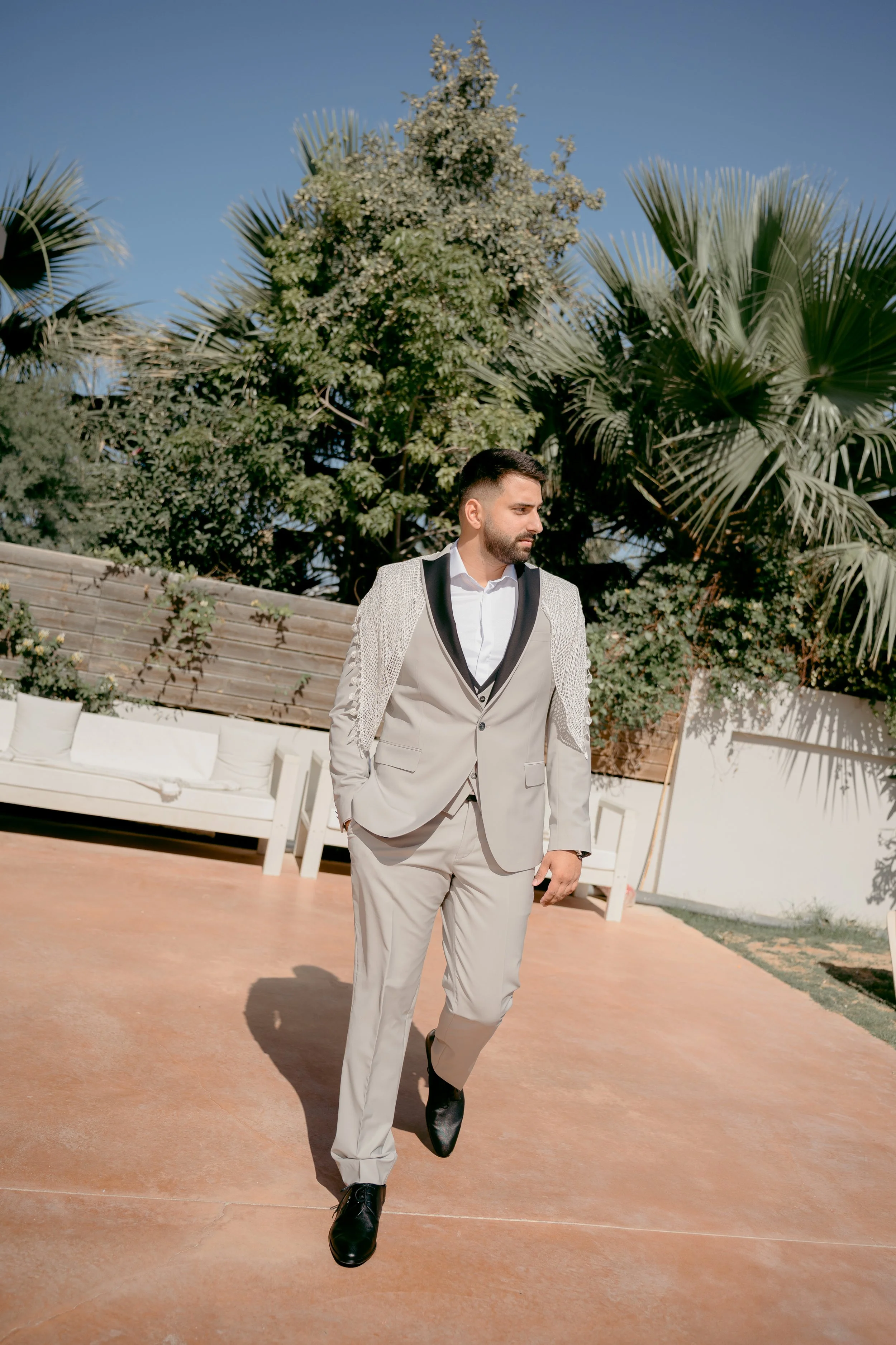 A man in a beige suit with black accents, white shirt, and black shoes walking outdoors on a sunny day. He has dark hair and a beard, with a garden background that includes trees and a lounge area with white cushions.