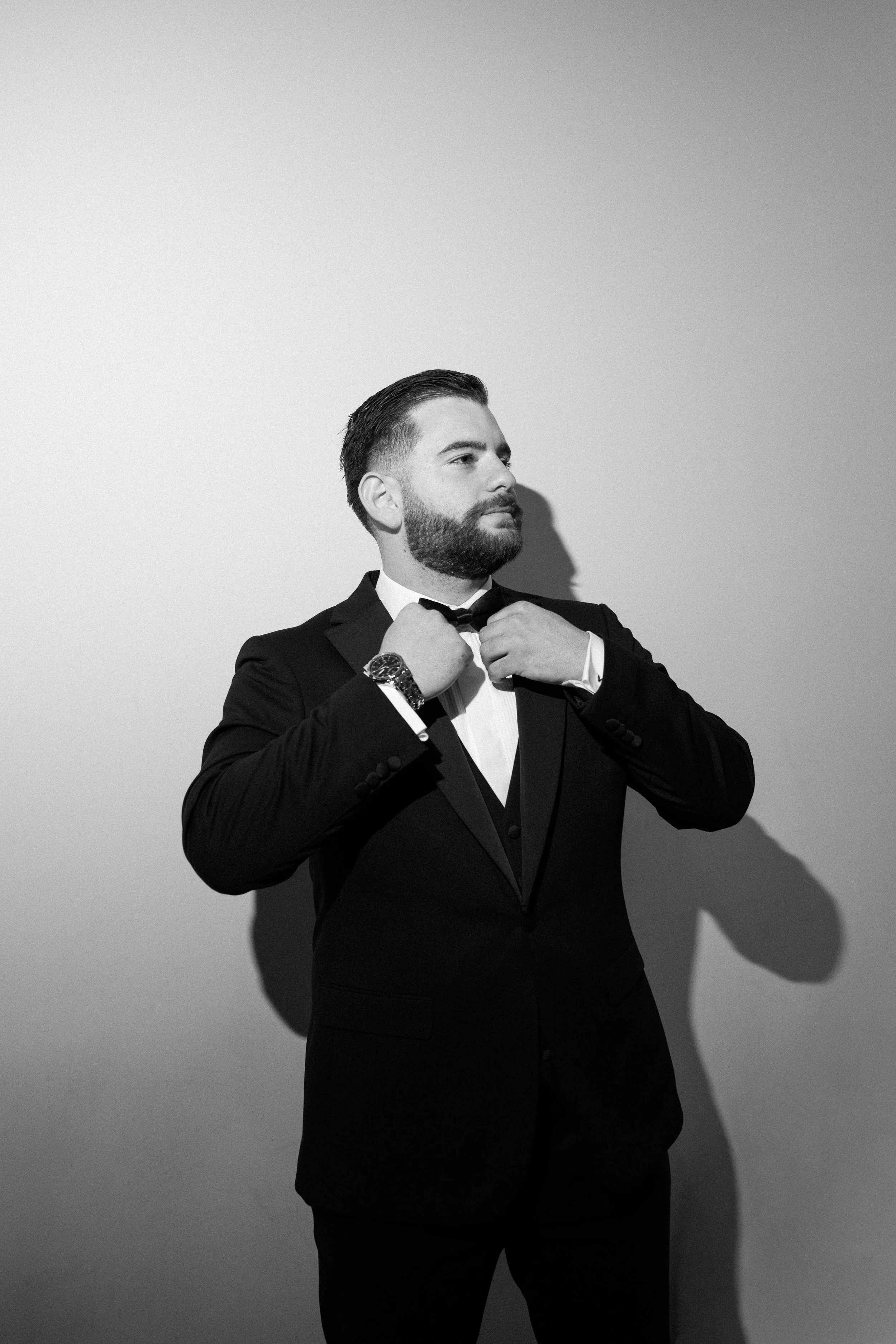 A man in a tuxedo adjusting his bowtie against a plain wall, casting a shadow, in black and white.