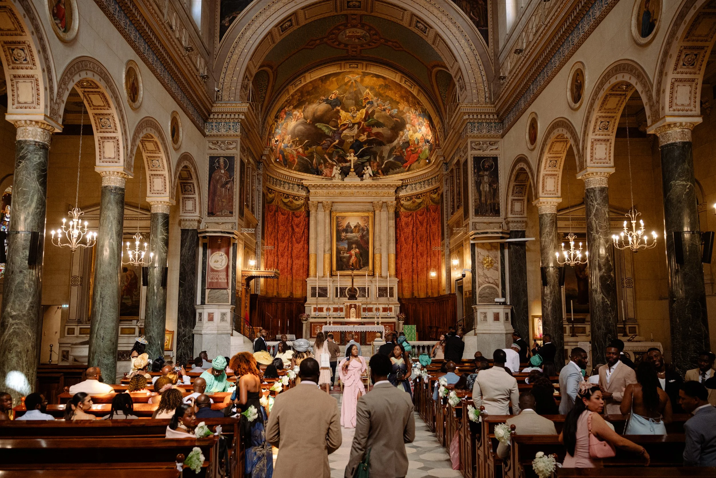 People attending a wedding ceremony inside a grand church or cathedral with ornate architecture and artwork, including paintings and a large ceiling mural, decorated with flowers and candles.