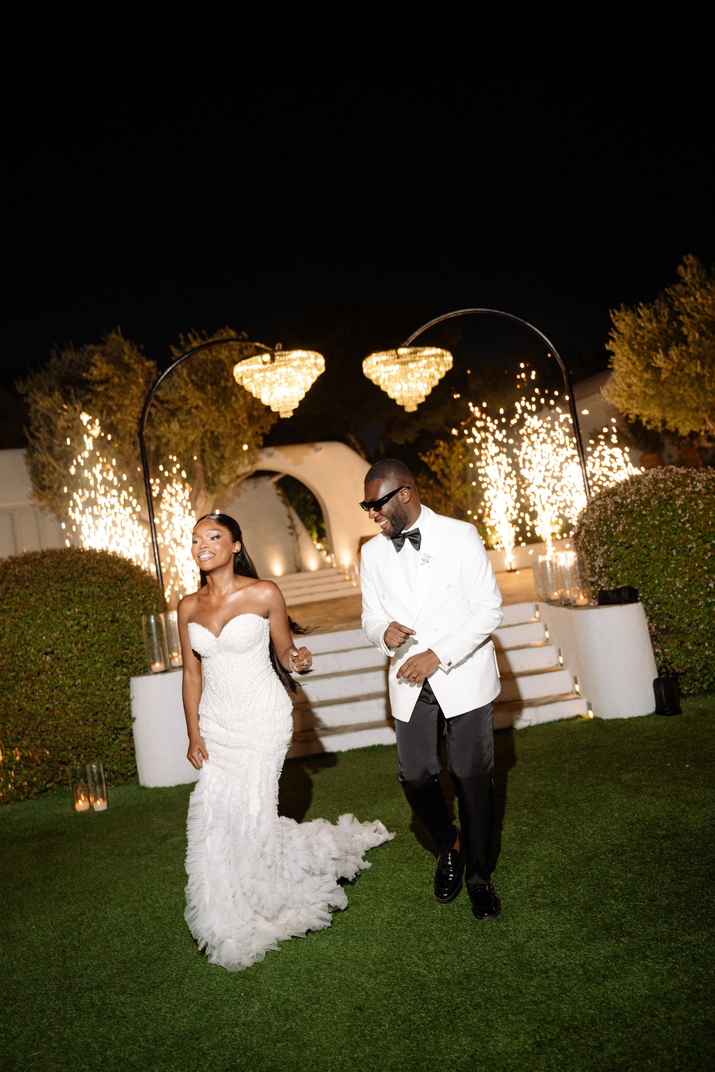 A newlywed couple dancing outdoors at night with fireworks and chandeliers illuminating the scene.