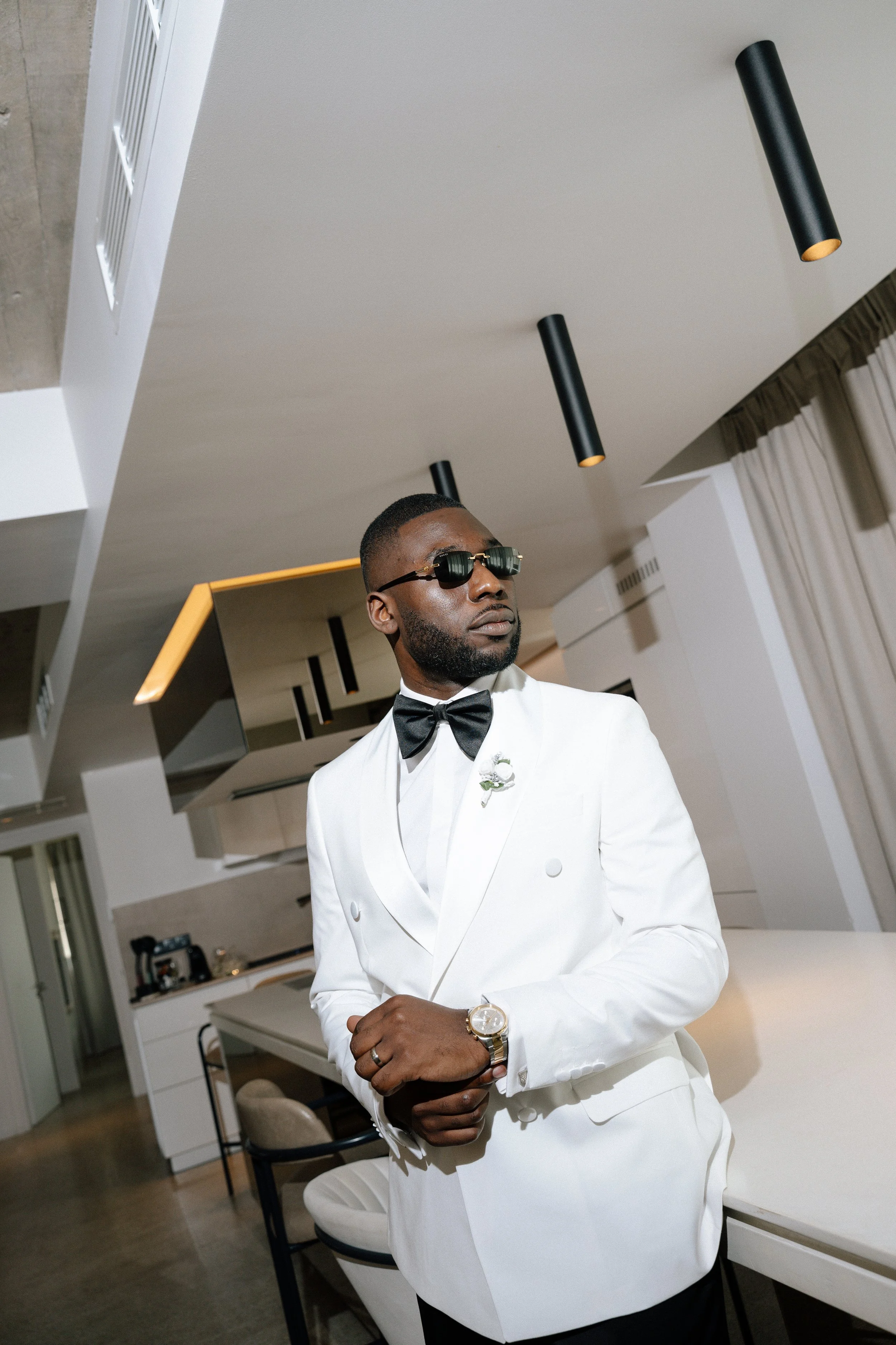 A man dressed in a white tuxedo with a black bow tie, wearing sunglasses and a watch, standing in a modern, well-lit room with a kitchen in the background.