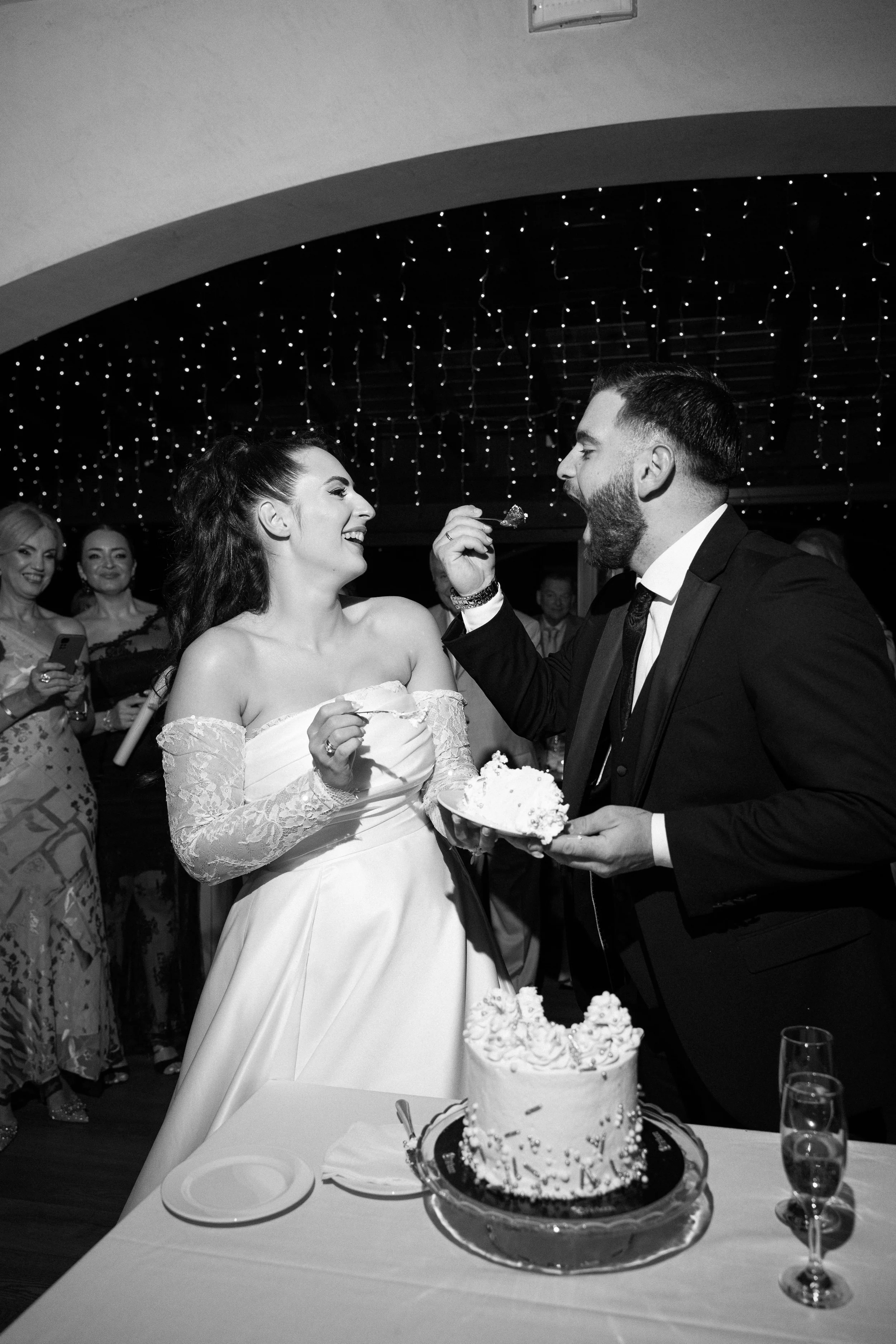 A bride and groom sharing cake at their wedding reception, with guests watching in the background.