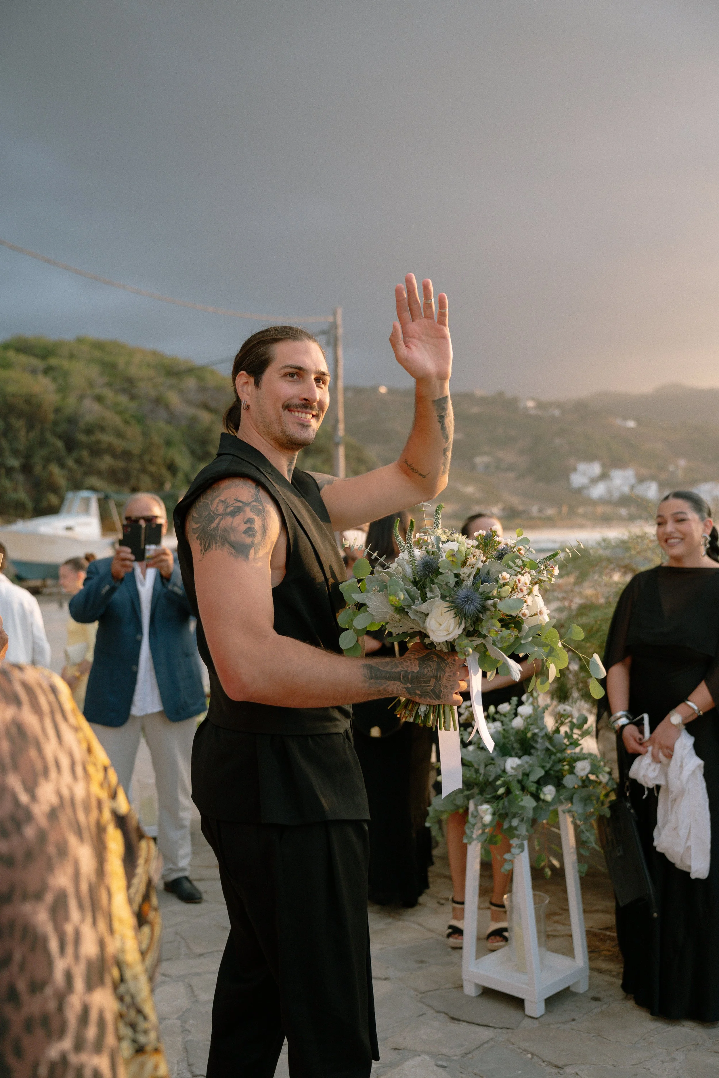 A man with tattoos on his arms, holding a bouquet of flowers, waving at an outdoor event during sunset, with a scenic hillside and a cloudy sky in the background.