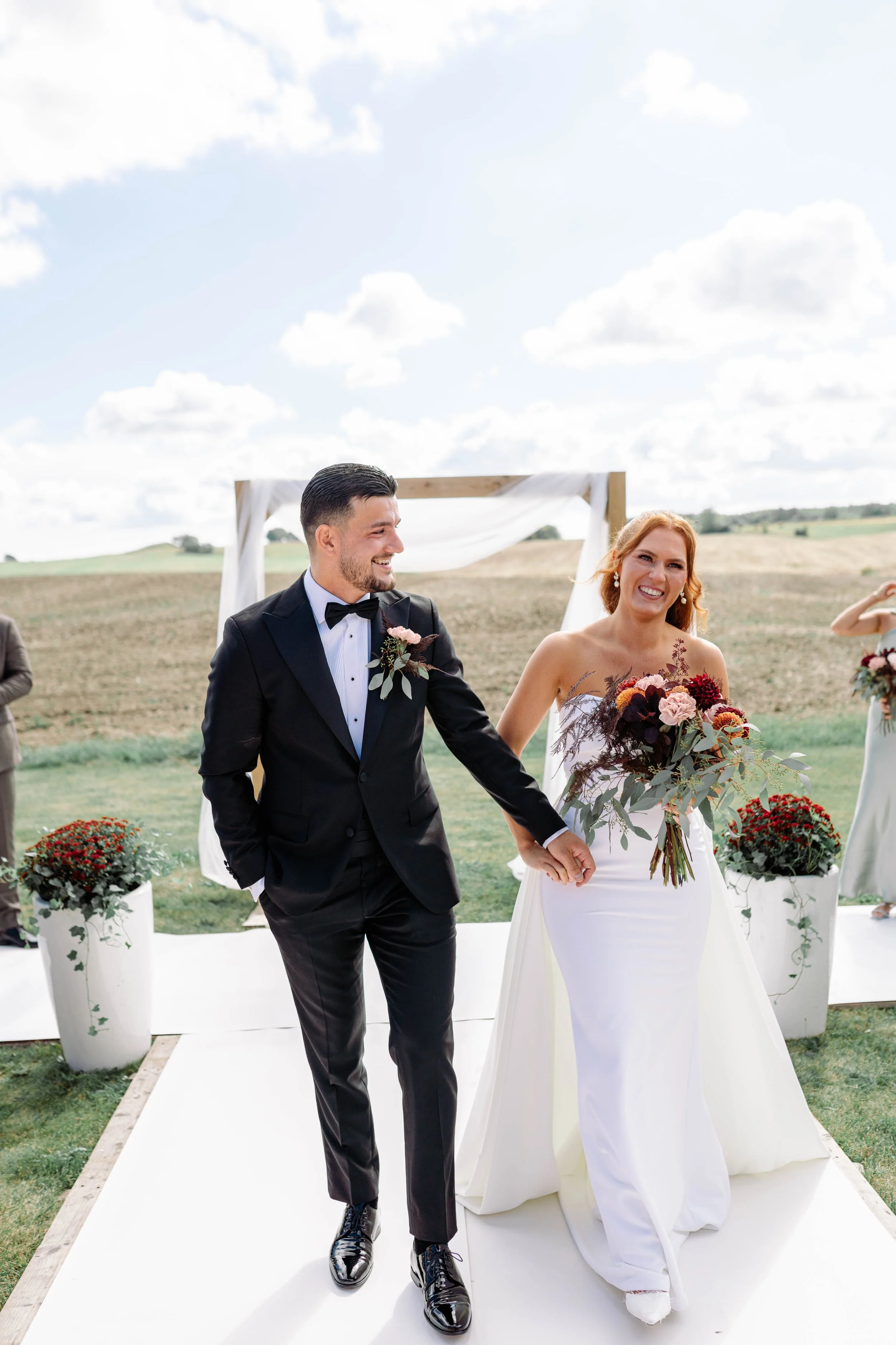 A newlywed couple walking outdoors after their wedding ceremony, holding hands and smiling, with a simple wooden arch and potted flowers in the background.