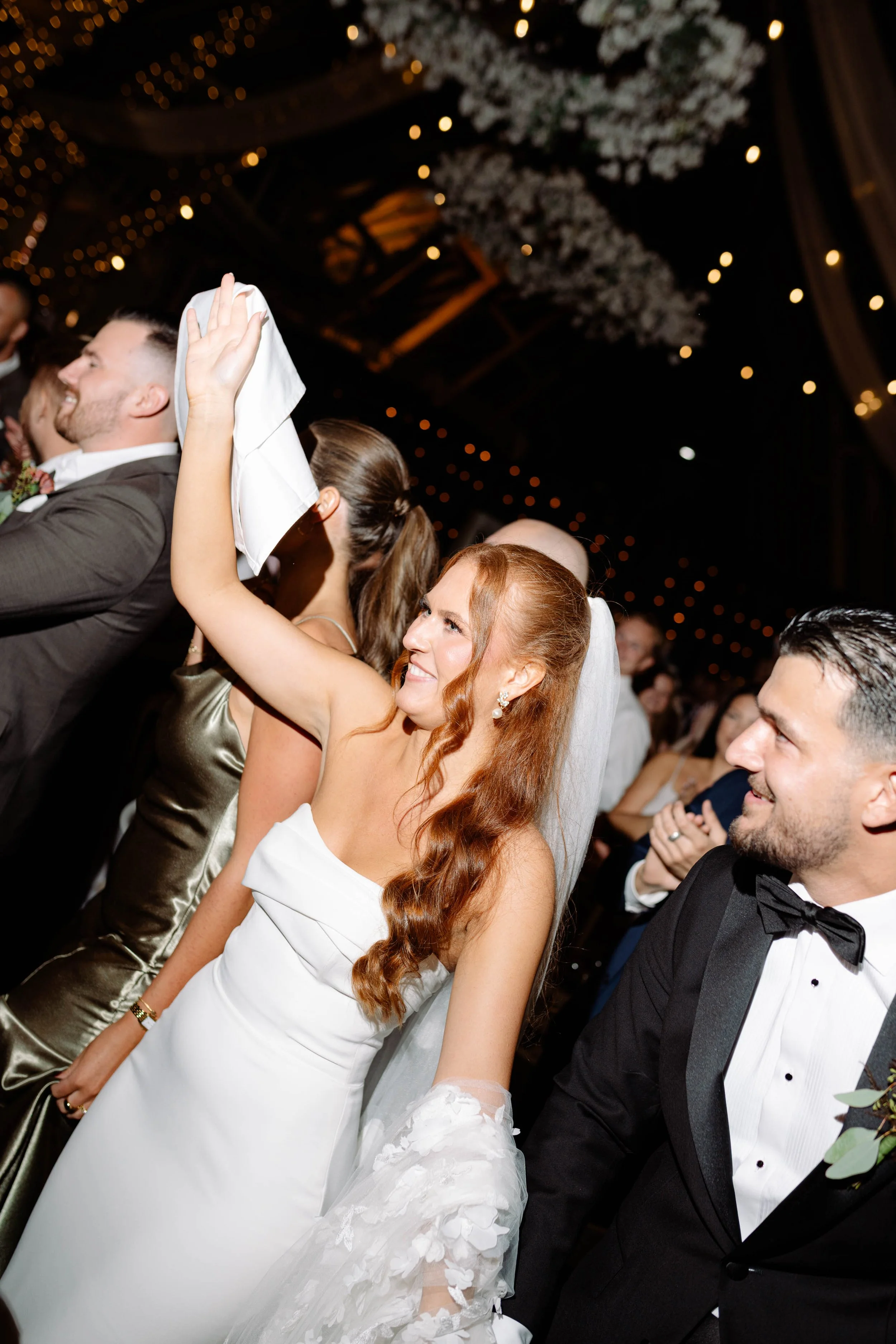 A woman with long red hair in a wedding dress, holding a white napkin, smiling, at a wedding celebration surrounded by other guests.