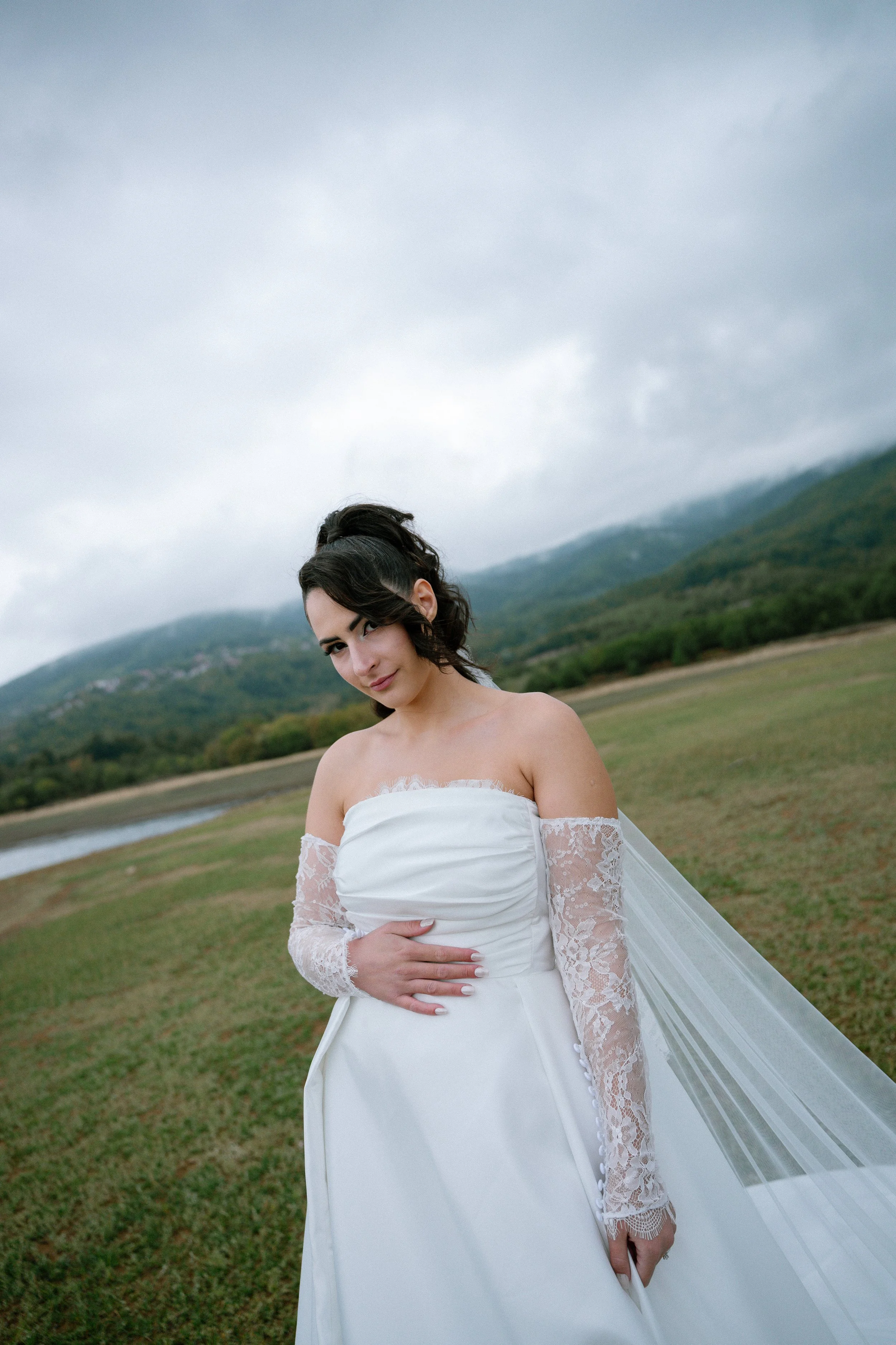 A woman in a wedding dress standing outdoors in a grassy field with mountains and cloudy sky in the background.