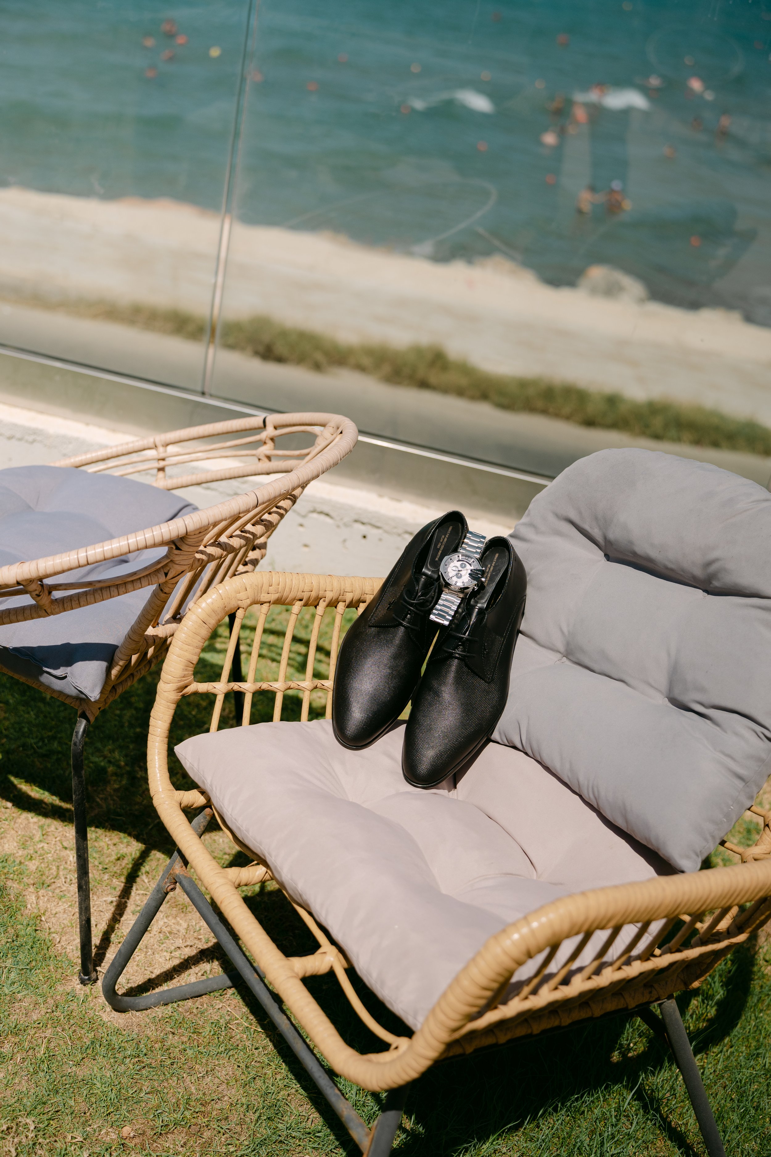 A wicker outdoor chair with a cushioned seat and a cushion on backrest, with black dress shoes and a silver watch placed on it, next to another wicker chair with a grey cushion, in front of a glass wall showing a sandy beach and water with swimmers.