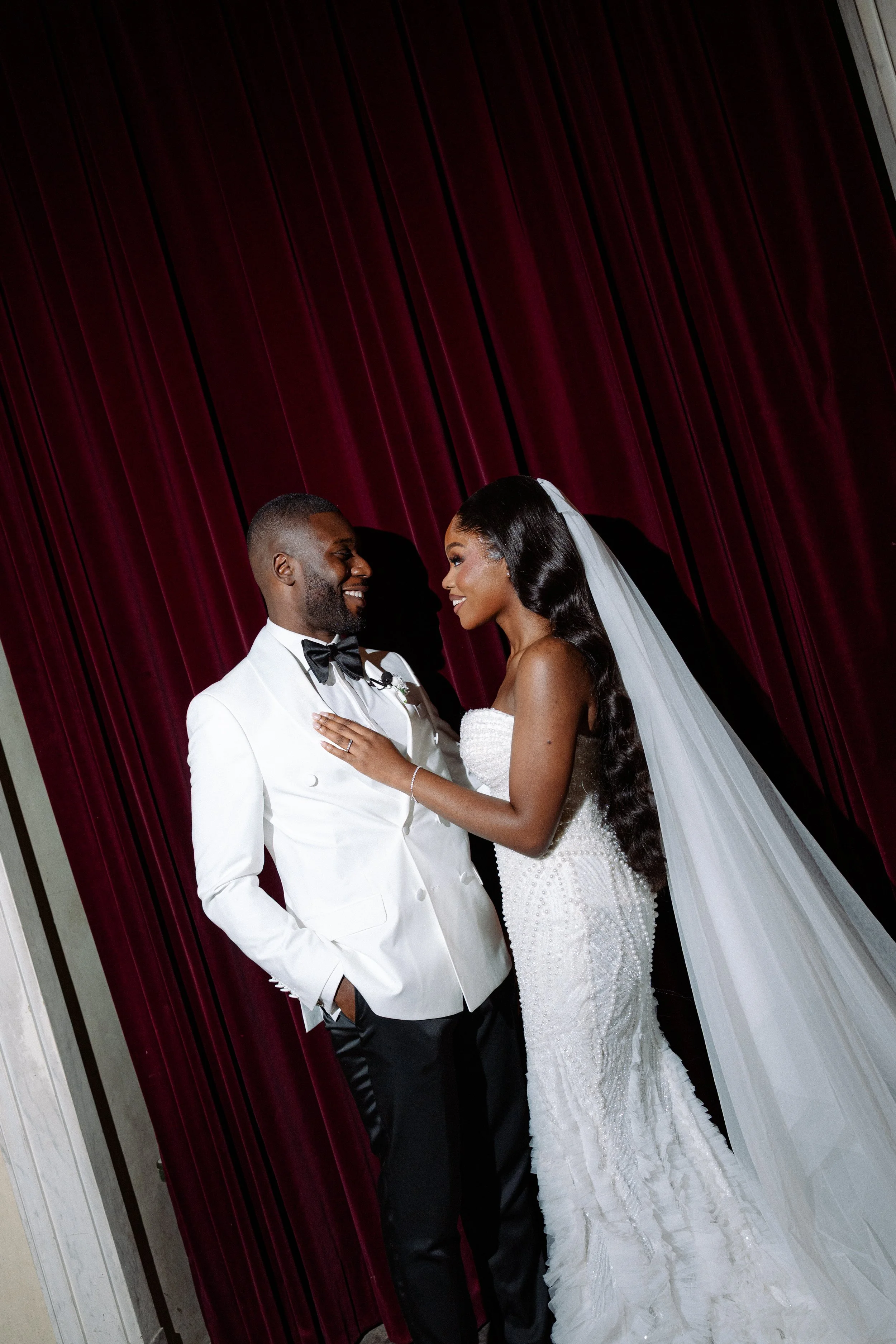 A bride and groom smiling at each other in front of dark red curtains, with the bride wearing a white wedding gown and veil, and the groom in a white tuxedo jacket and black bow tie.