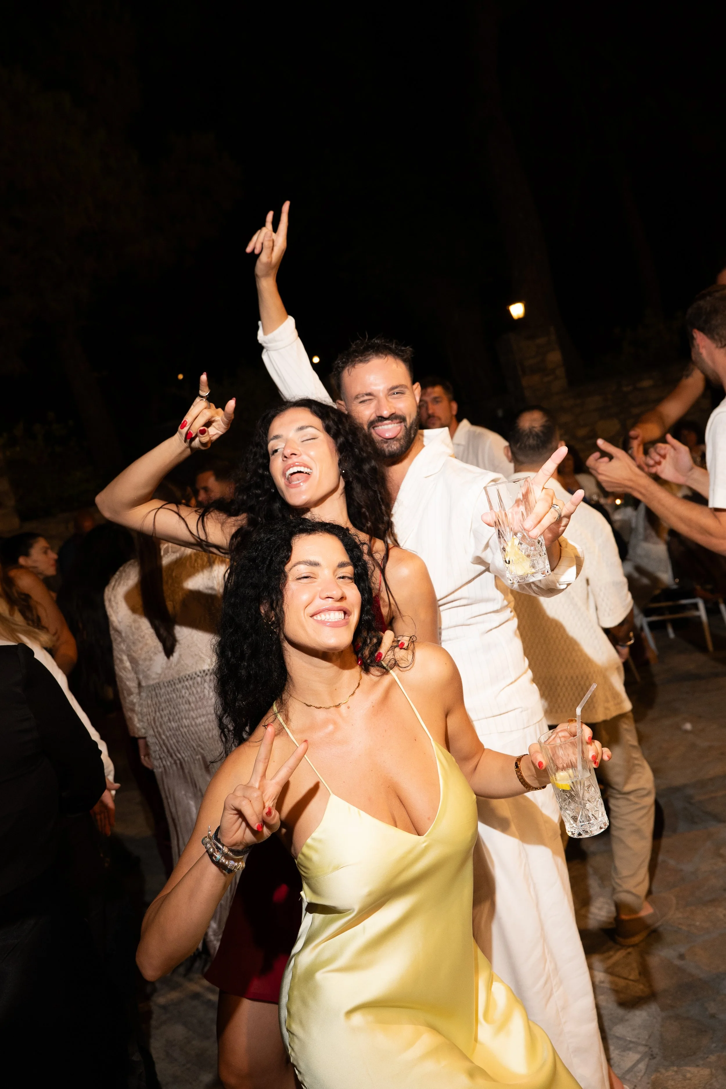 Three people celebrating at a party, holding drinks and making peace signs, with others dancing in the background.