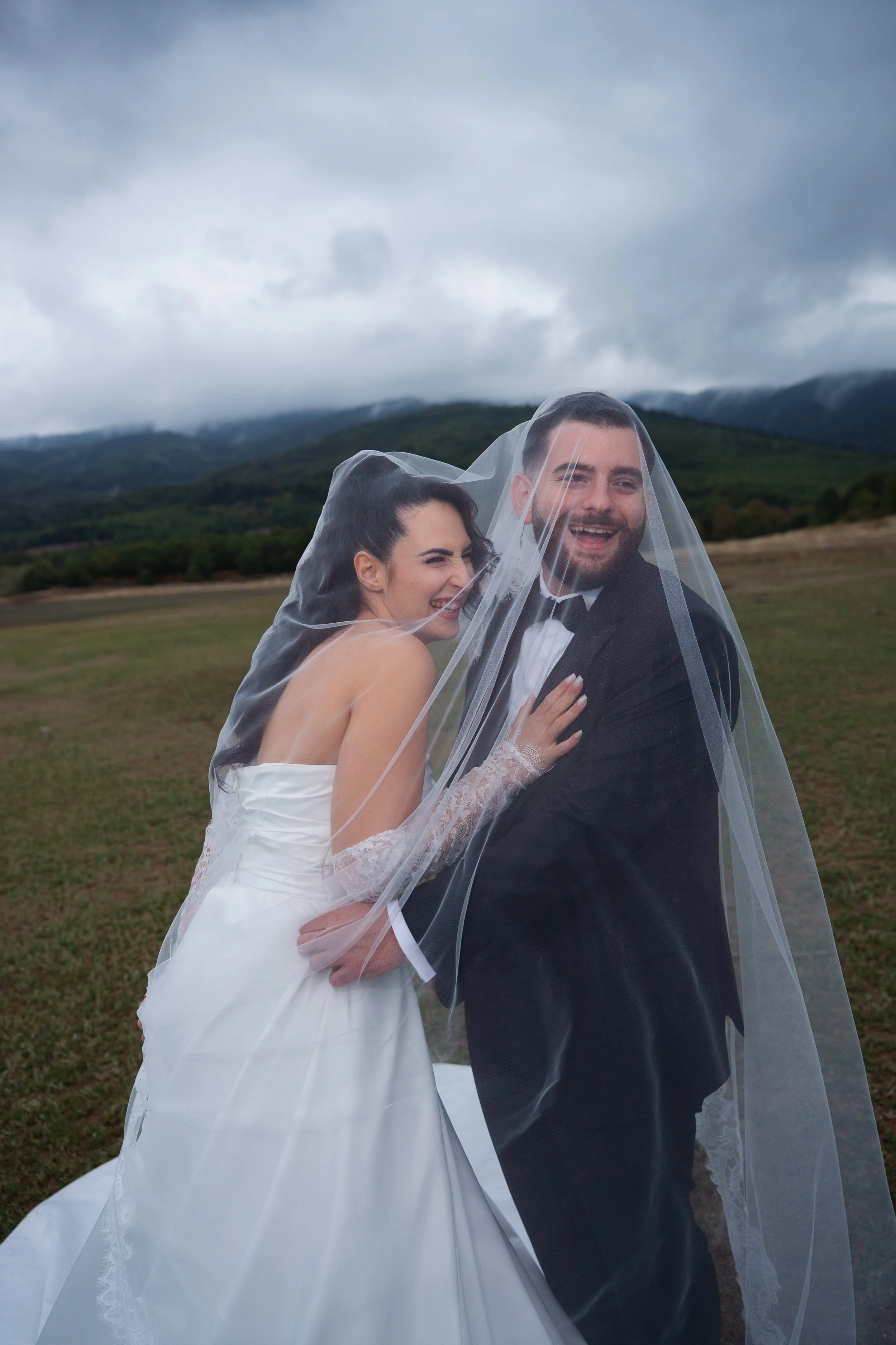 Bride and groom under a veil, smiling, standing outdoors with mountains and cloudy sky in the background.