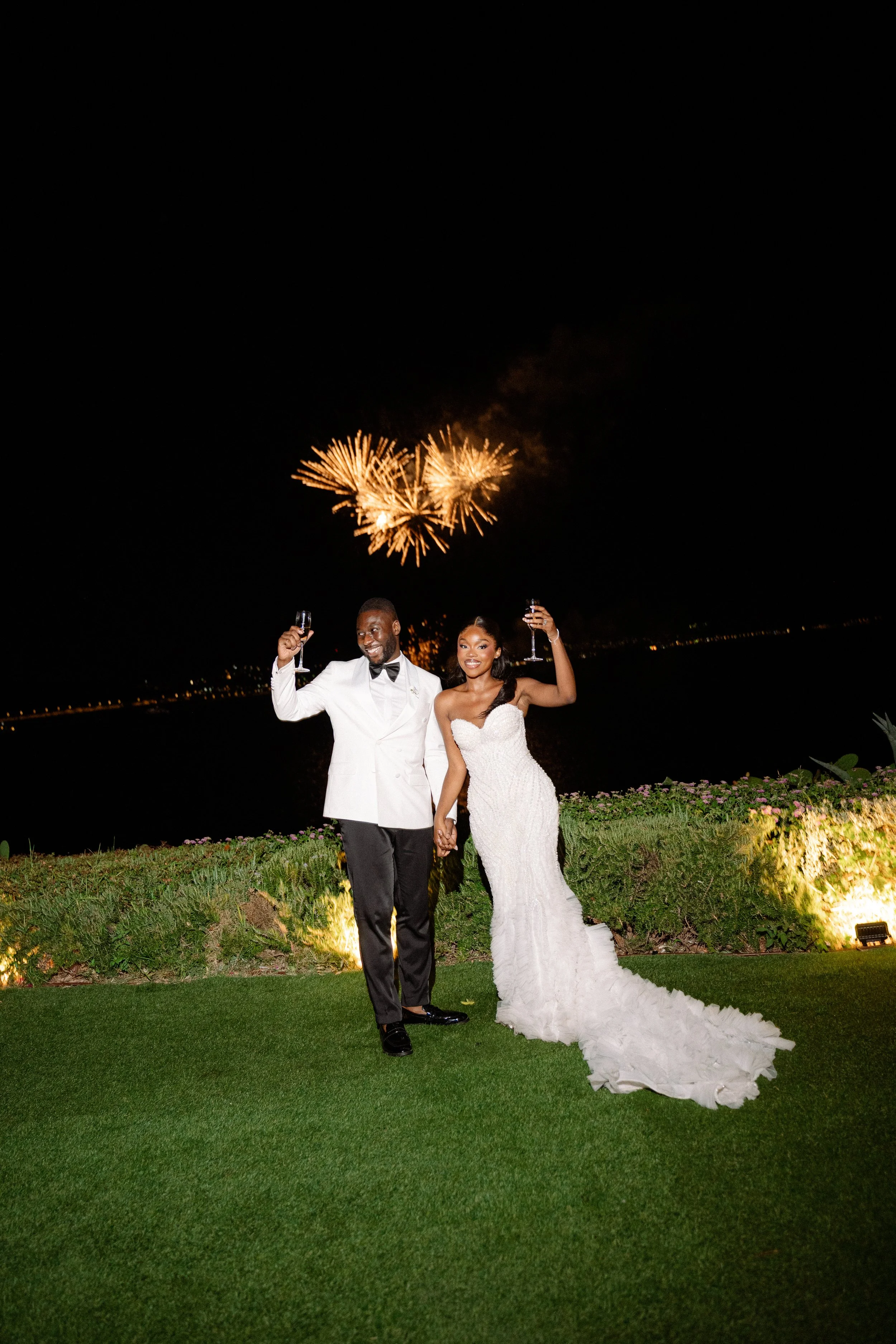 A newlywed couple celebrating with drinks at night with fireworks in the sky behind them, standing on a grassy area with flowers.