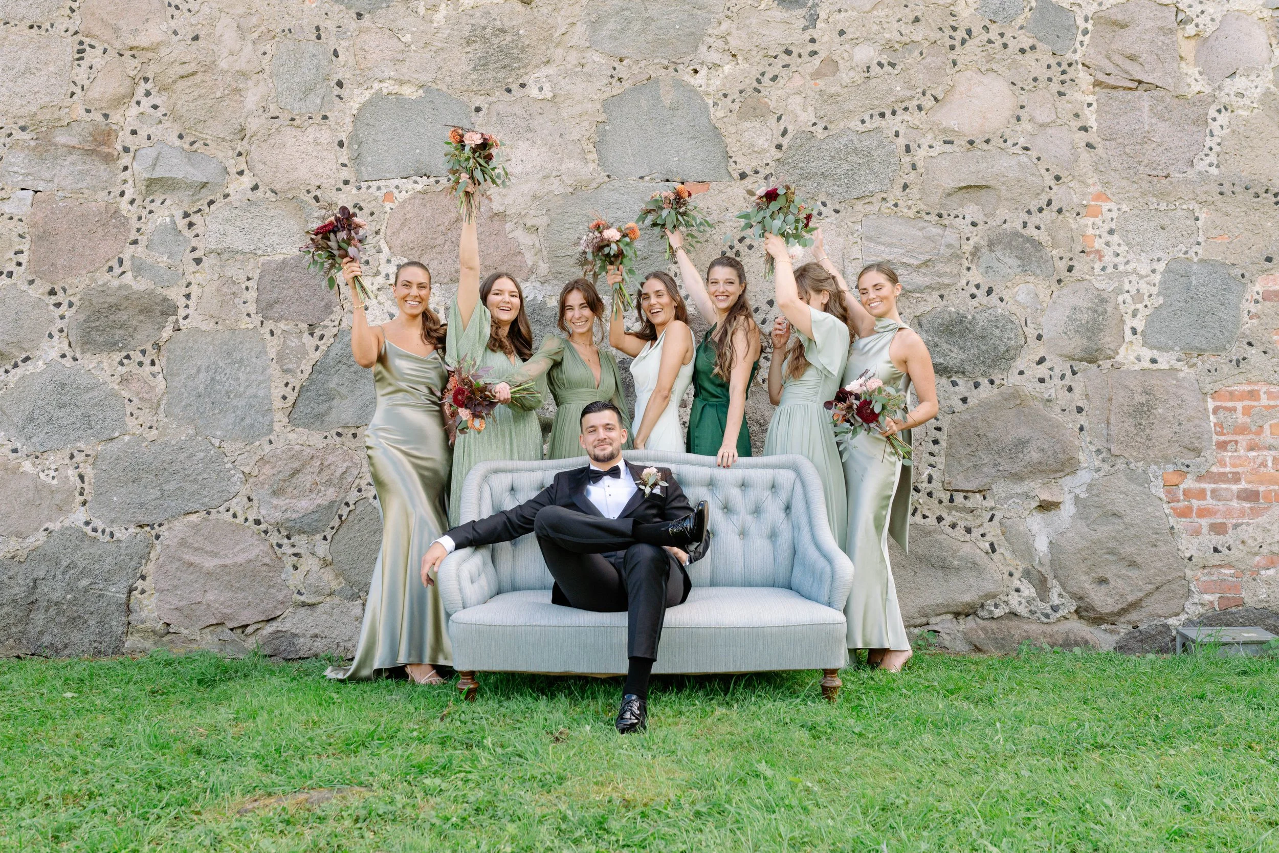 Group of women in elegant dresses and a man in a tuxedo, posing outdoors against a stone wall, with some women holding bouquets and raising their arms in celebration.