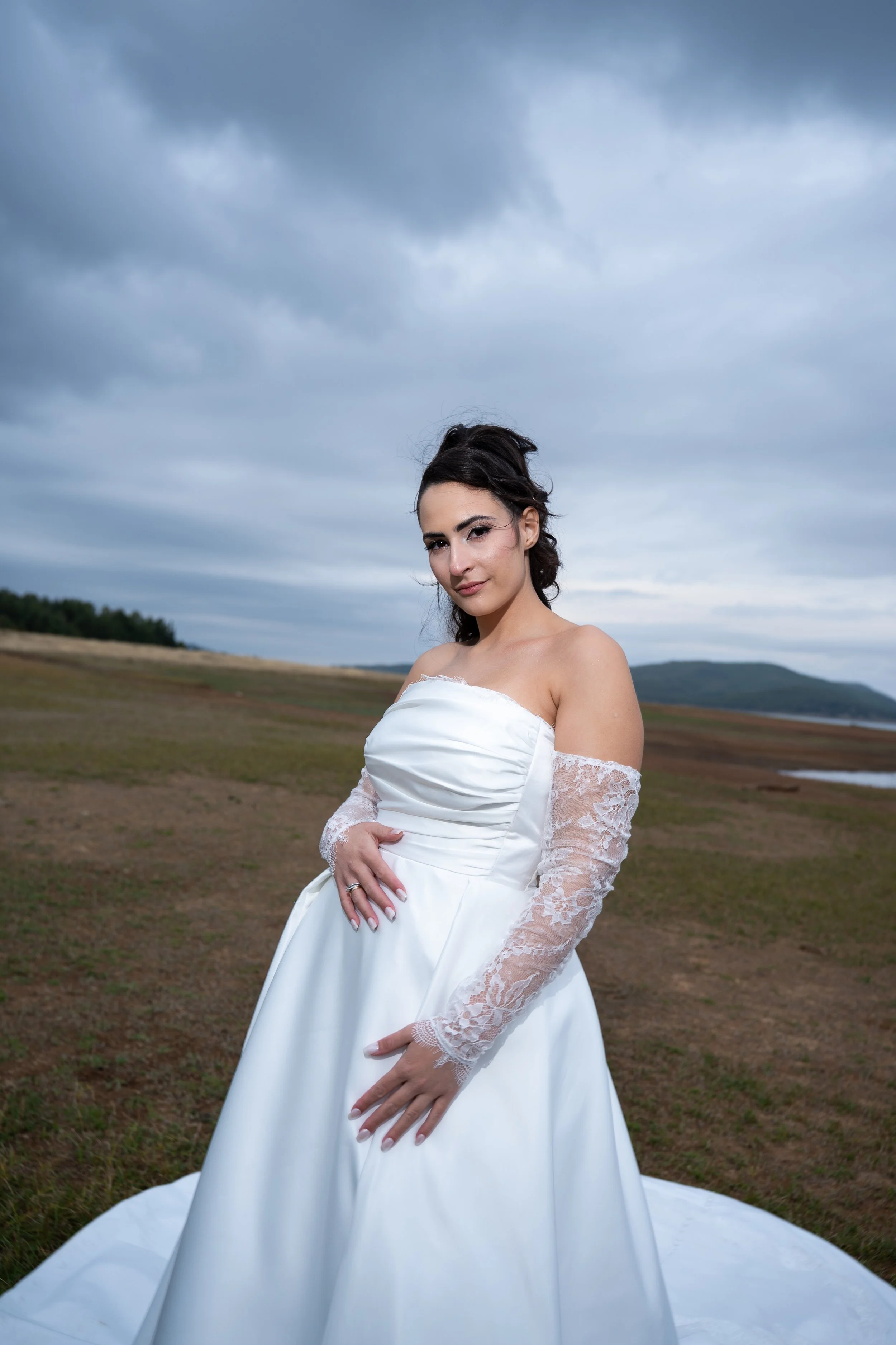 A woman in a white wedding dress with lace sleeves standing outdoors on a cloudy day.