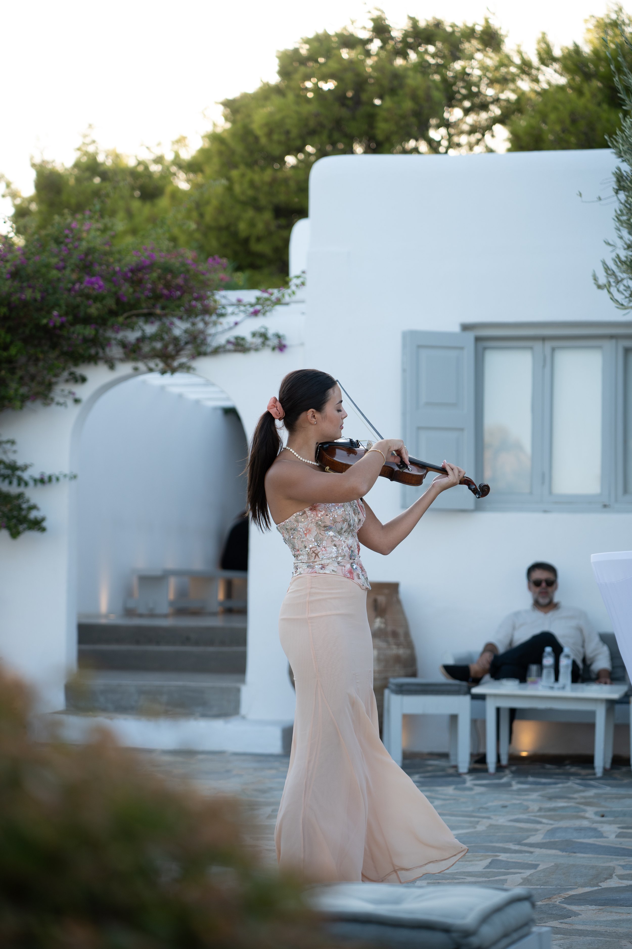 A woman in a strapless dress playing the violin outdoors in a Mediterranean-style setting with white walls and greenery, while a man in sunglasses relaxes on a bench in the background.