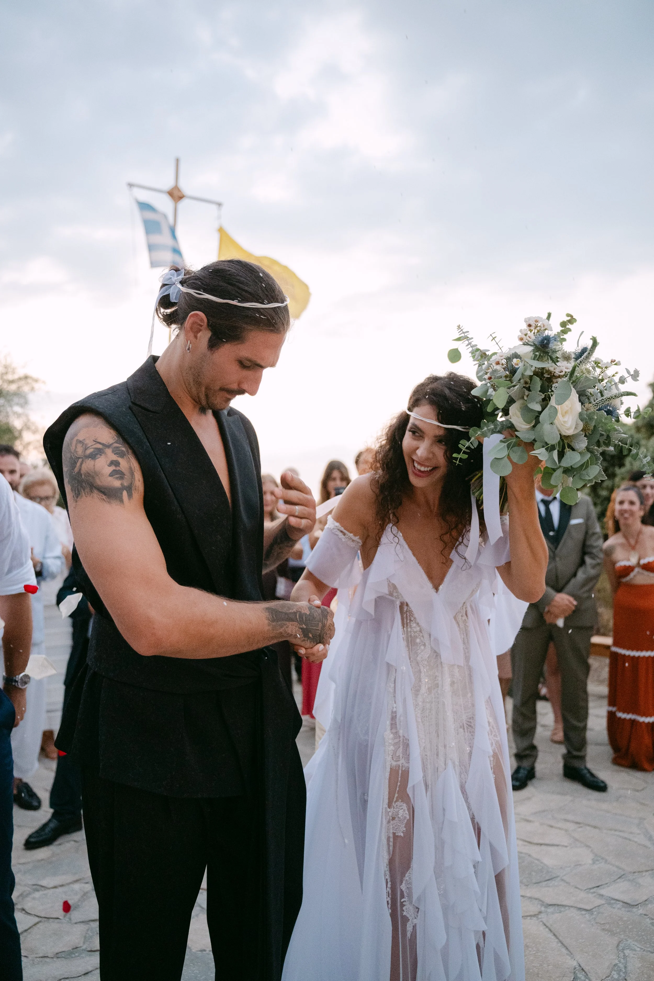 A couple getting married outdoors, with a woman in a white dress holding a bouquet and a man in black attire with tattoos. They are surrounded by guests and flags in the background, during sunset.