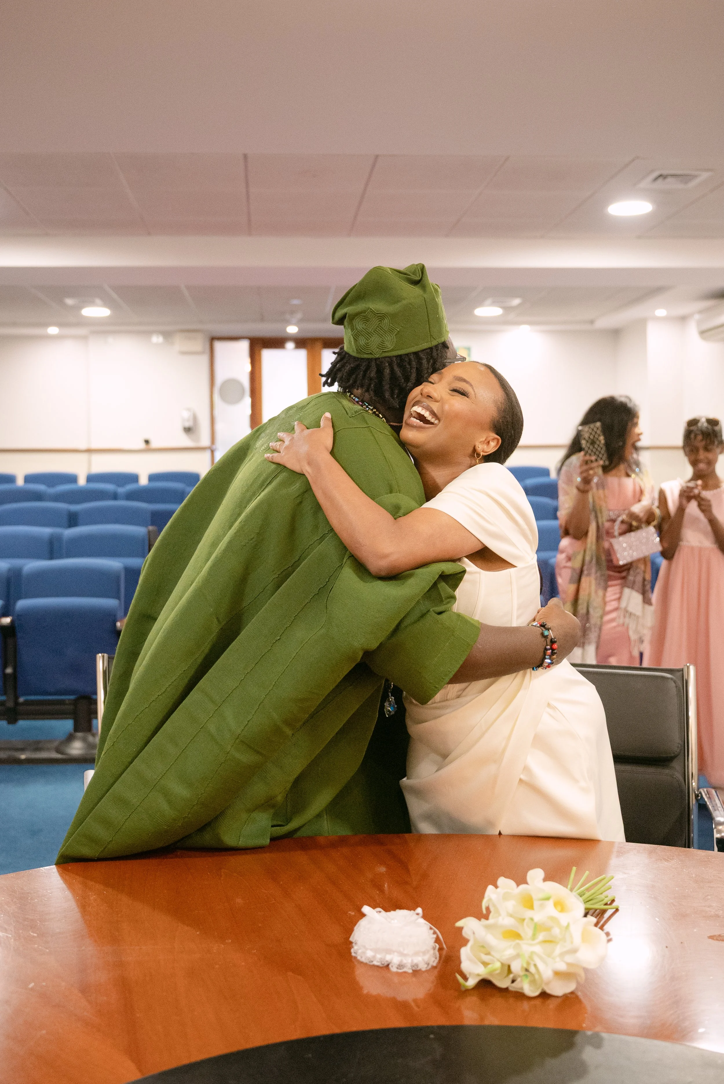 Two women hugging and smiling happily in a room with blue chairs, with three other women in the background taking photos and wearing light pink dresses.