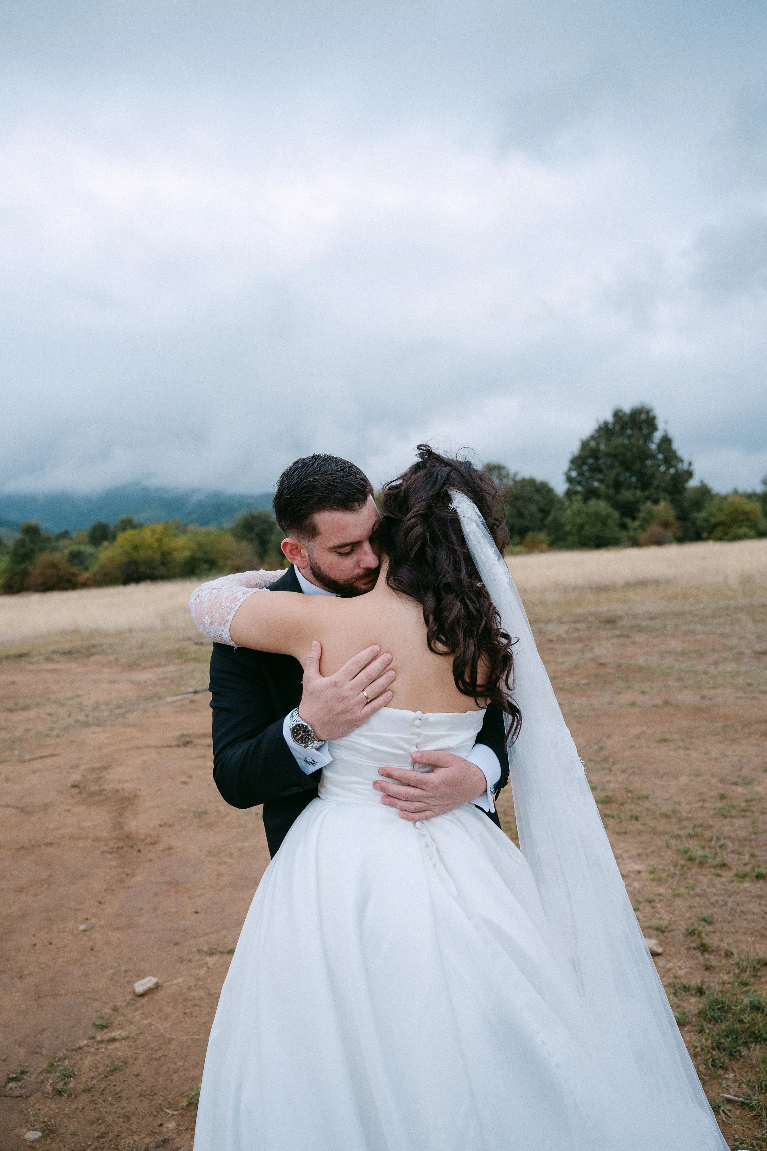 A bride and groom share a hug in an outdoor setting, with the bride wearing a white wedding dress and veil and the groom in a dark suit, against a backdrop of cloudy sky, trees, and open field.