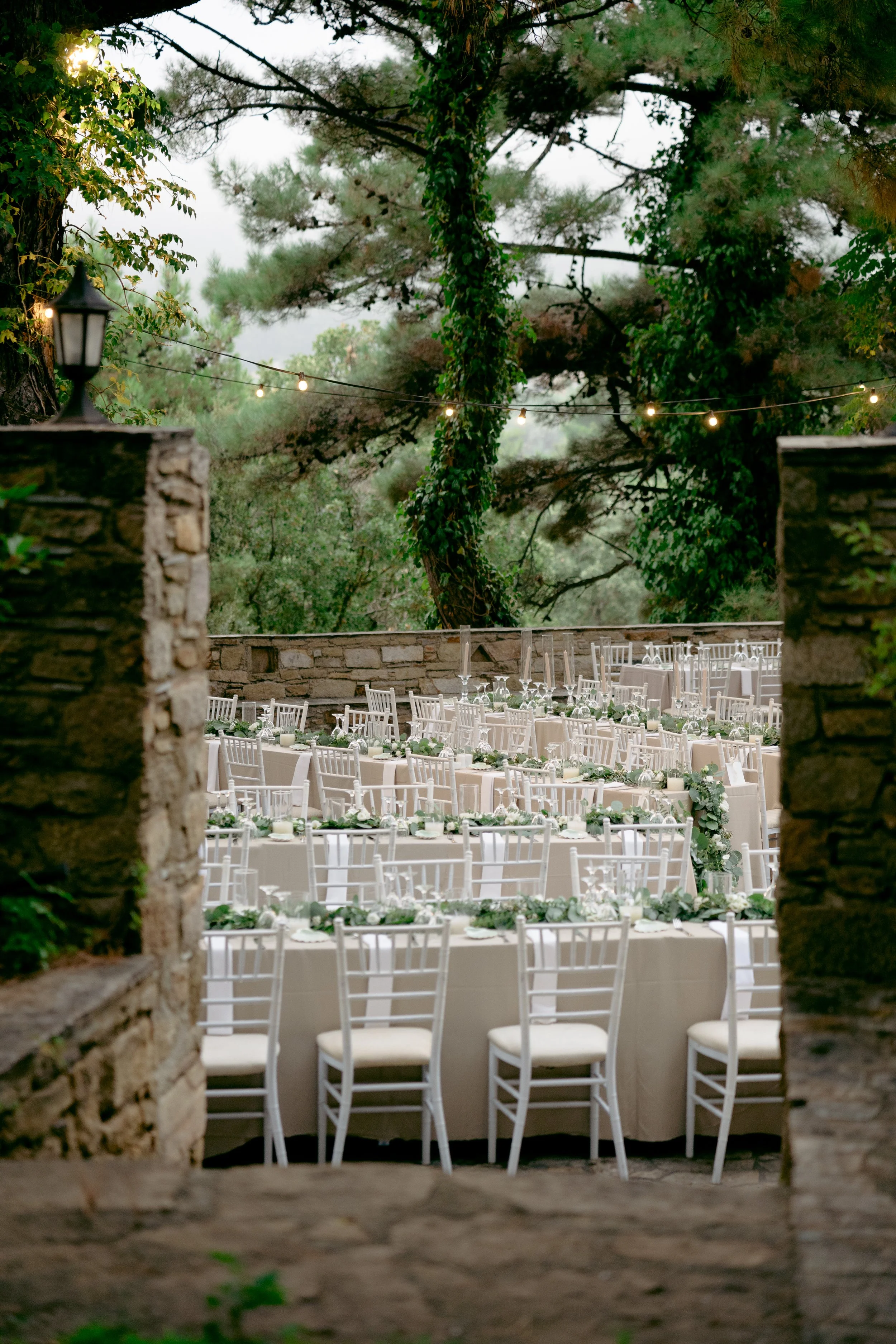 Outdoor wedding reception setup with long tables, white chairs, and greenery decorations, surrounded by trees and stone walls.