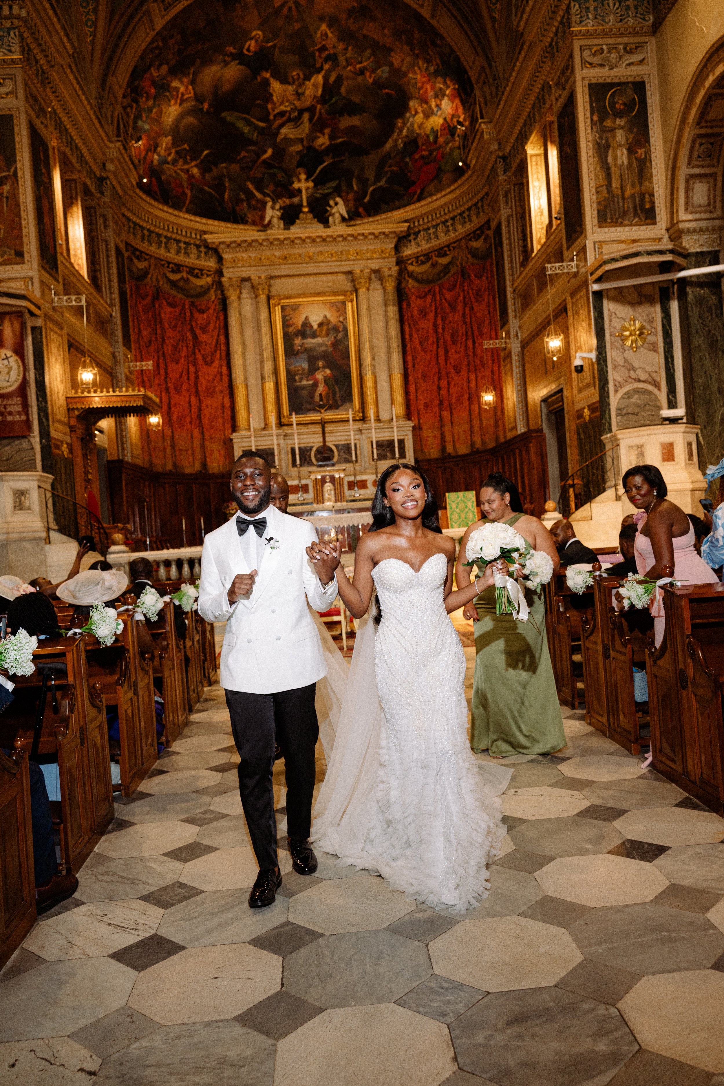 Bride and groom walking down the aisle in a church, smiling, with wedding guests seated on either side, ornate decorations, and a large religious painting and artwork on the ceiling.