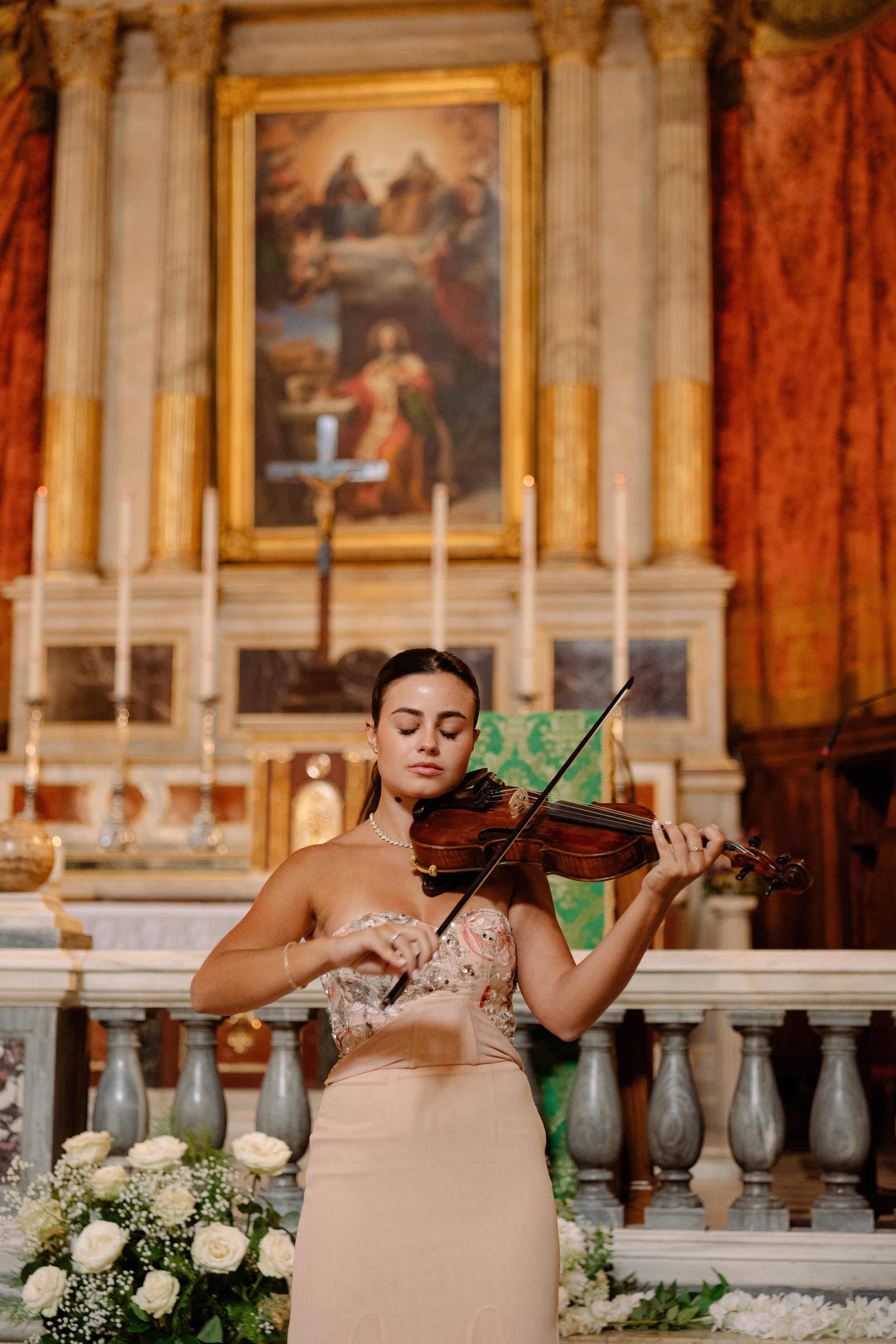 Young woman in a strapless dress playing the violin in a church with floral arrangements and a large religious painting in the background.
