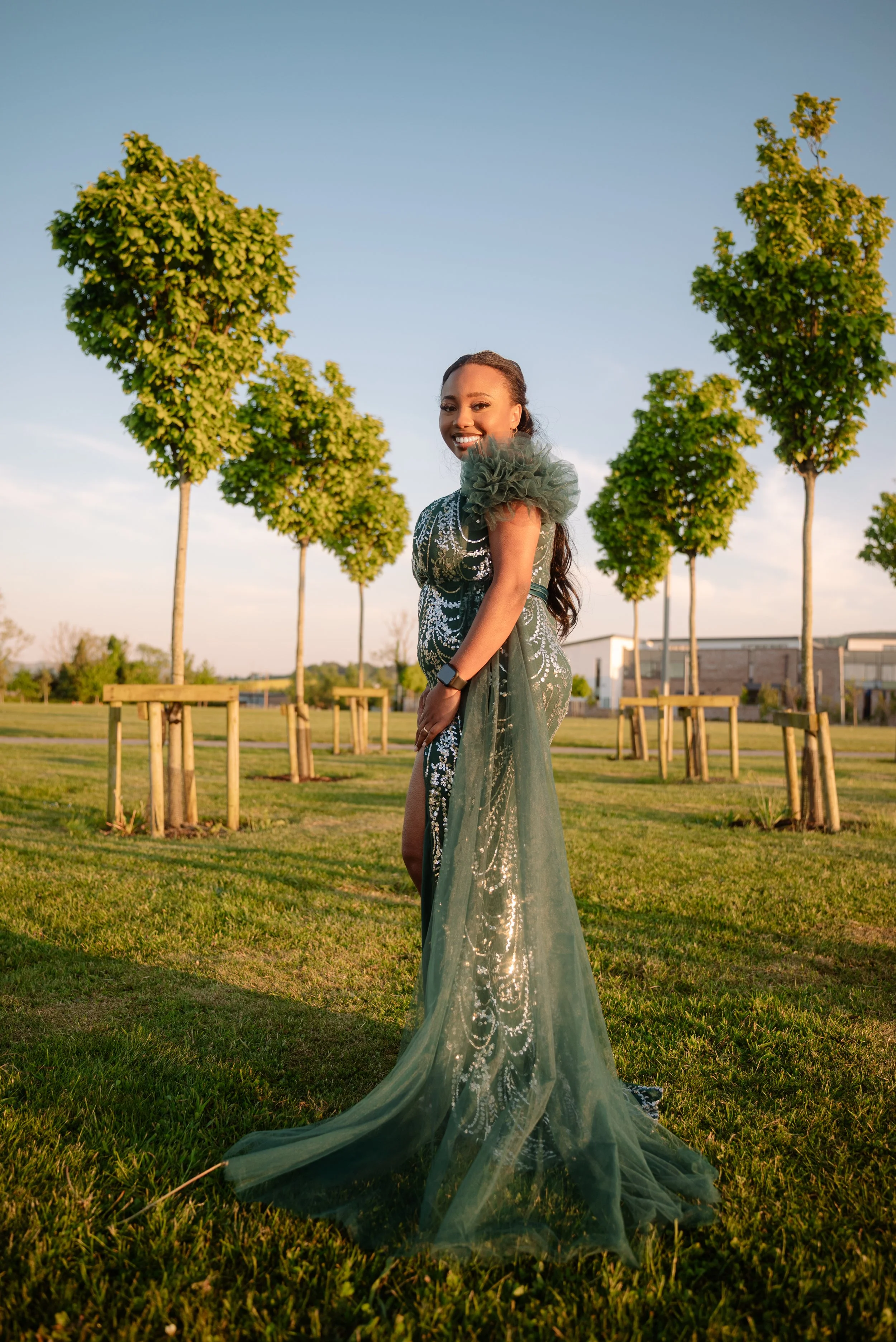 A woman in a glamorous green dress with tulle and sequin details standing outdoors on a sunny day, smiling, with small trees and a building in the background.