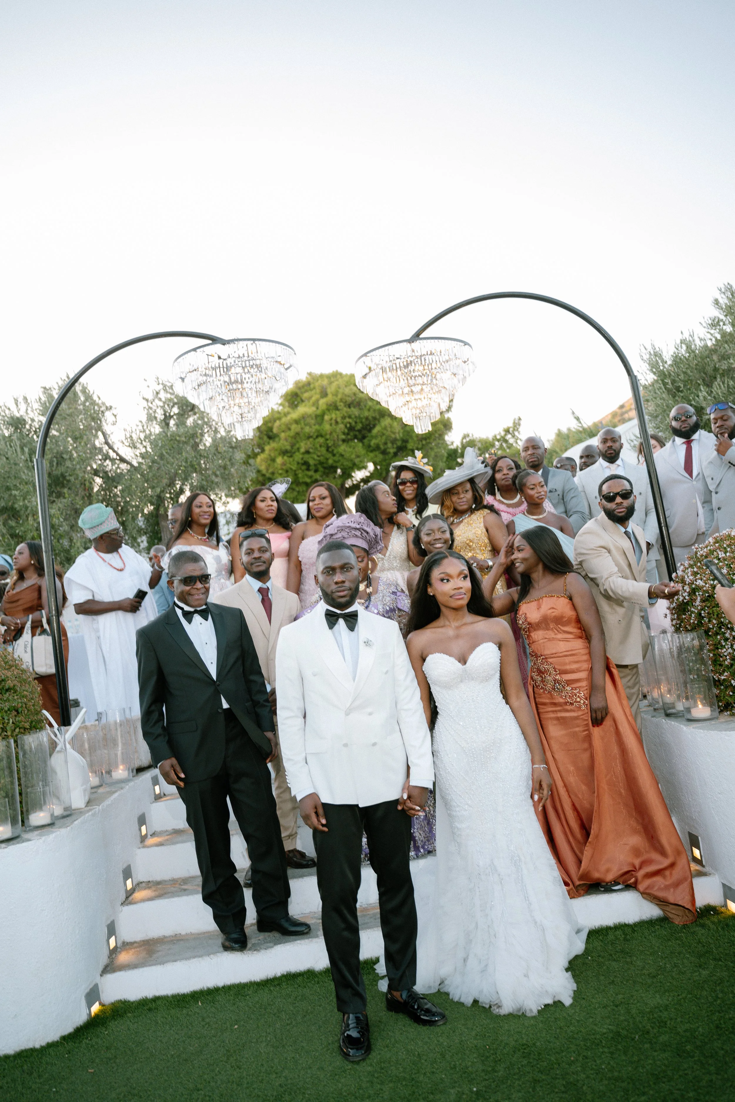 A large wedding party group photo outdoors with the bride and groom in front, surrounded by family and friends, under decorative lighting and a tree, during evening.