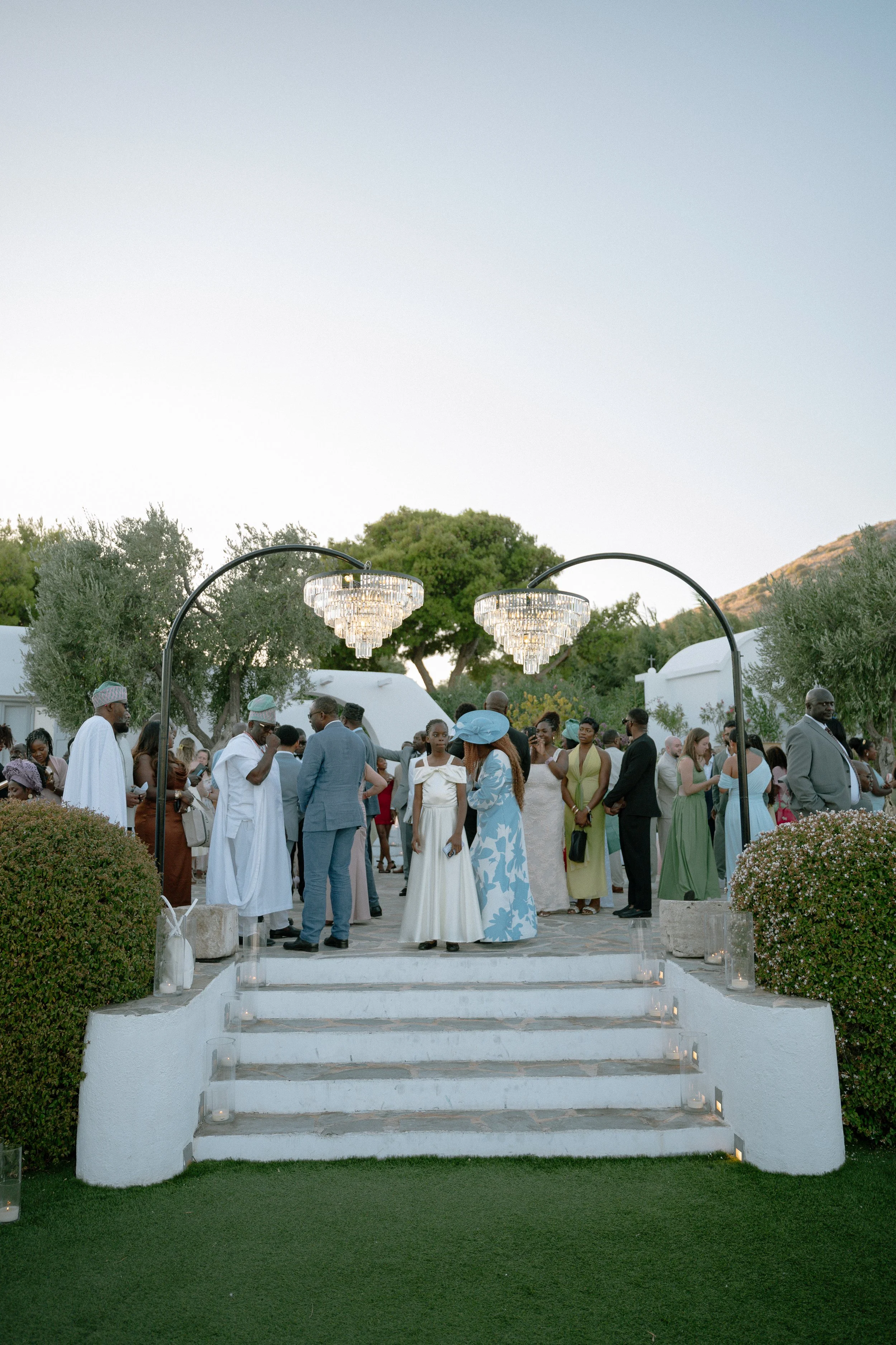 People gathered at an outdoor wedding ceremony during the daytime, standing on a white stone platform with steps, surrounded by greenery and trees.