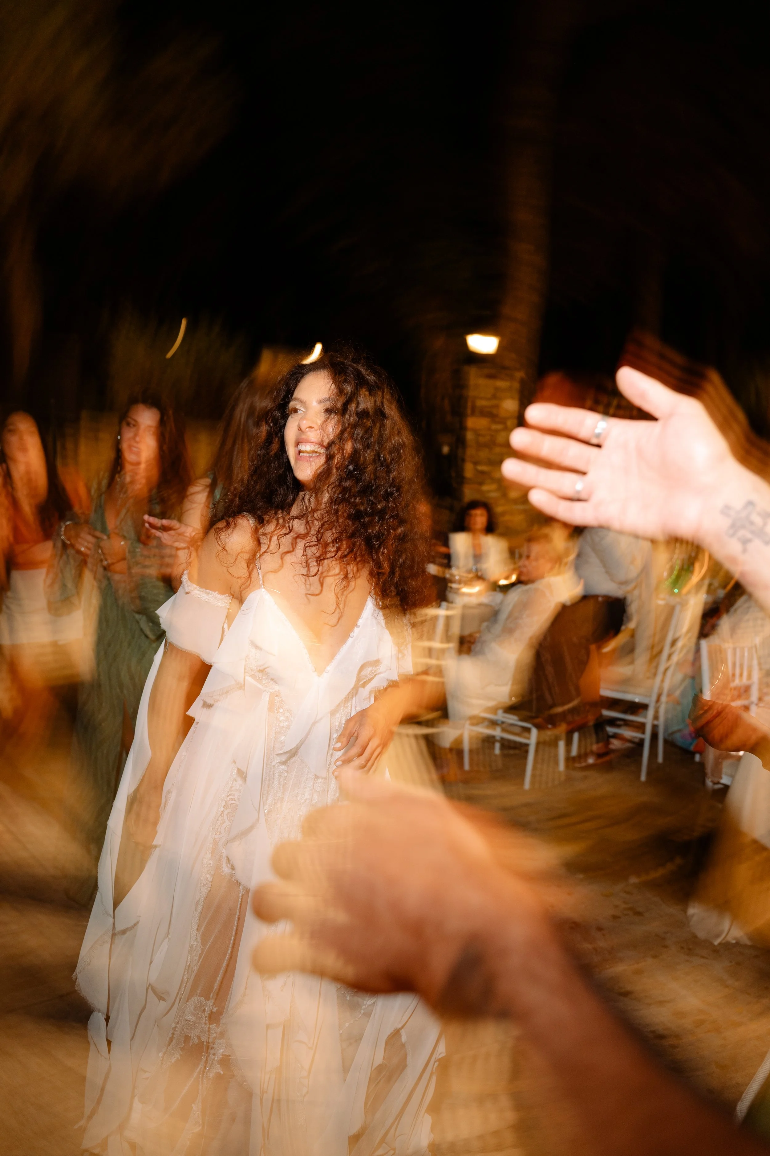 A woman with curly hair dancing and smiling at a lively party or celebration. Other guests are visible in the background, and the setting appears to be indoors with warm lighting.