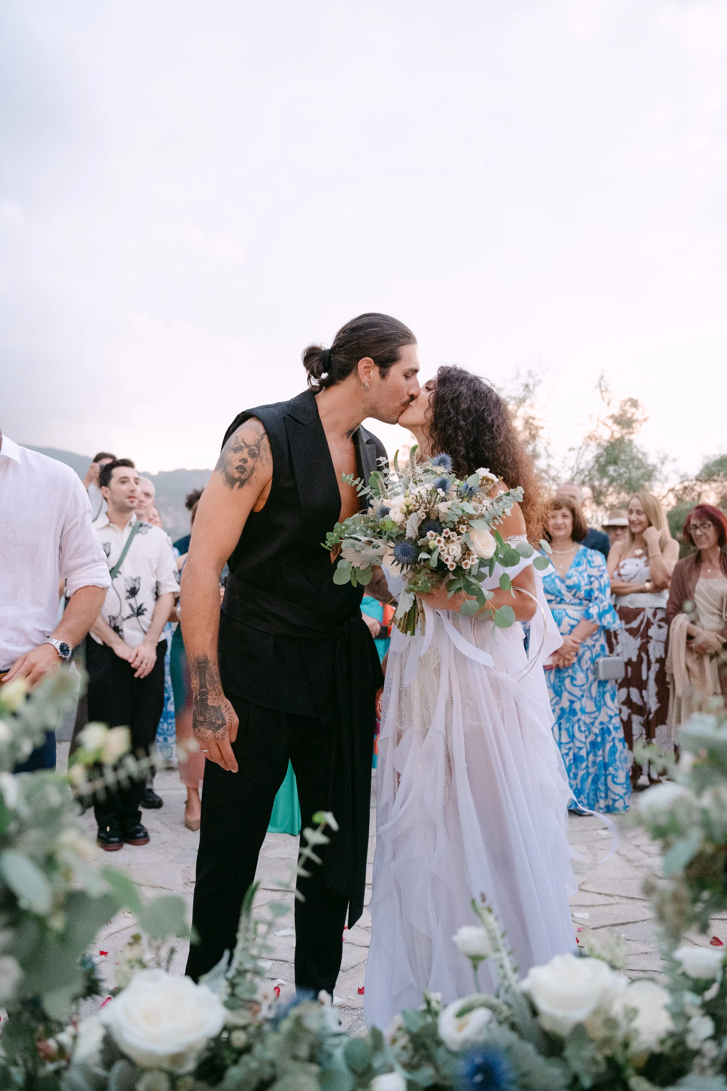 A same-sex couple kisses during their outdoor wedding ceremony at sunset, with guests watching in the background.
