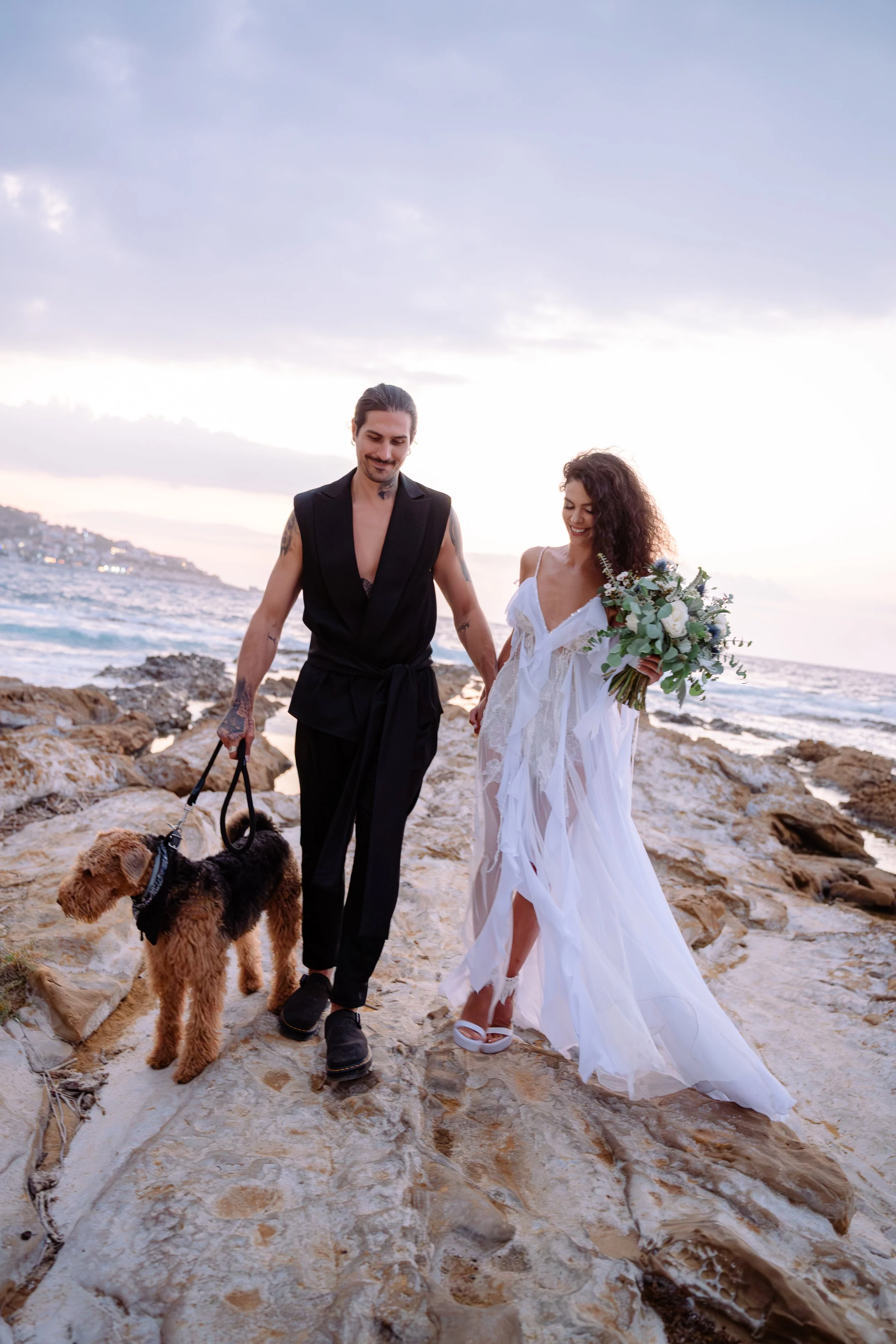 A couple walking on rocky beach at sunset, holding hands, with a small dog, ocean in the background, the woman wearing a white flowing dress and holding a bouquet of flowers.