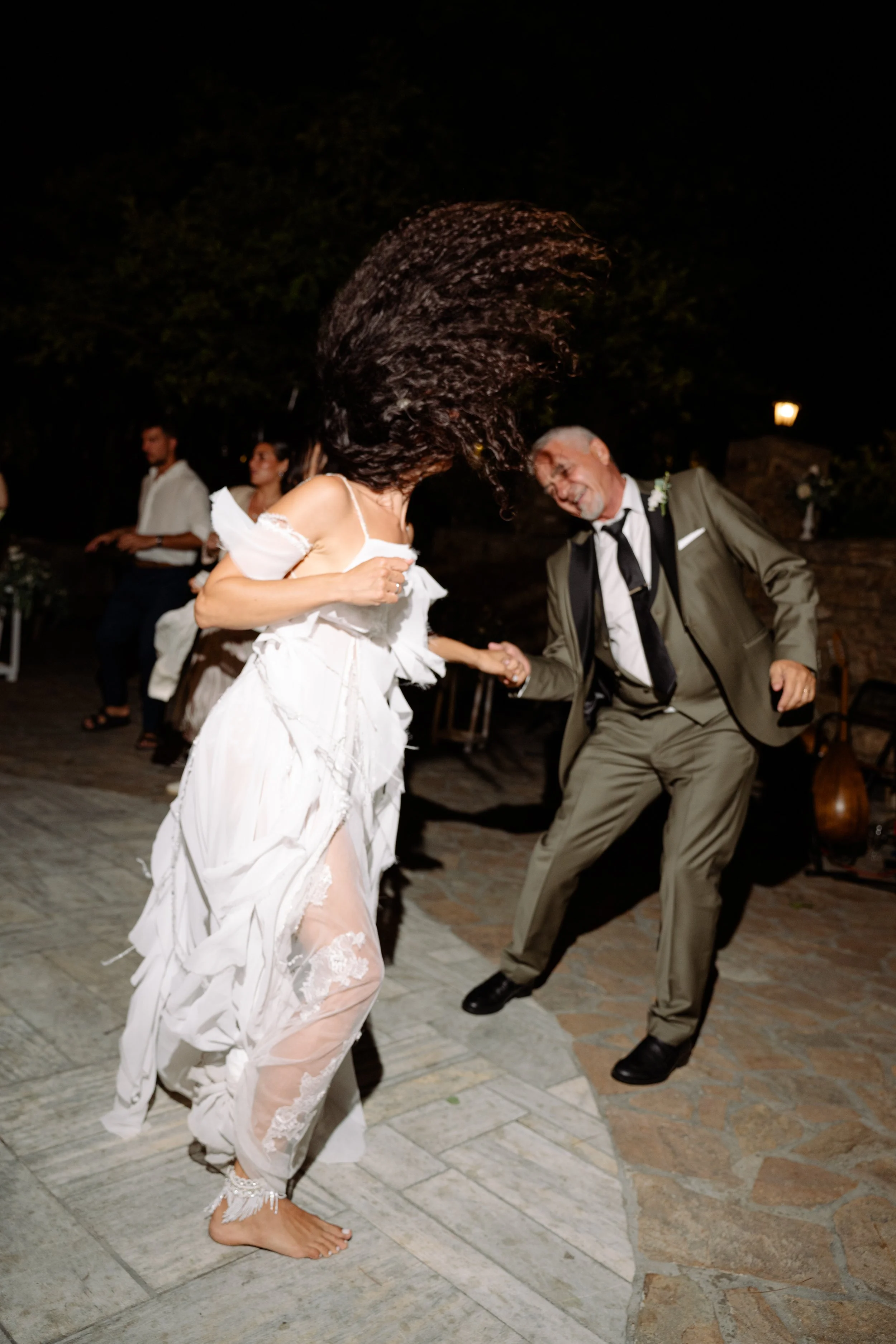 Two people are dancing at an outdoor event at night. The woman is wearing a white dress and the man is in a gray suit. They are holding hands and moving energetically, with the woman's hair flying through the air.
