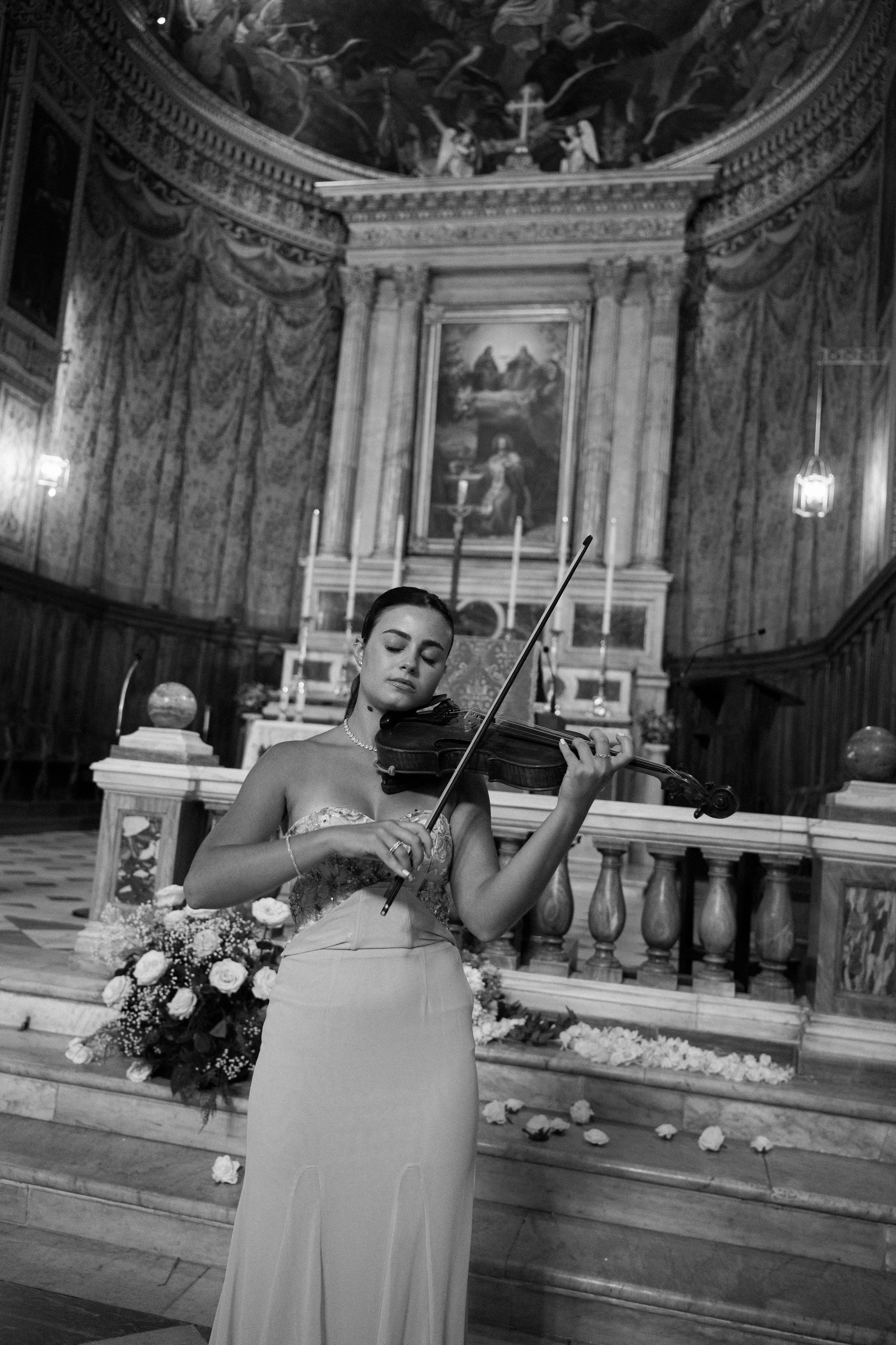 A young woman in a strapless gown playing the violin inside a church with an ornate altar and religious paintings, and flowers on the stairs.