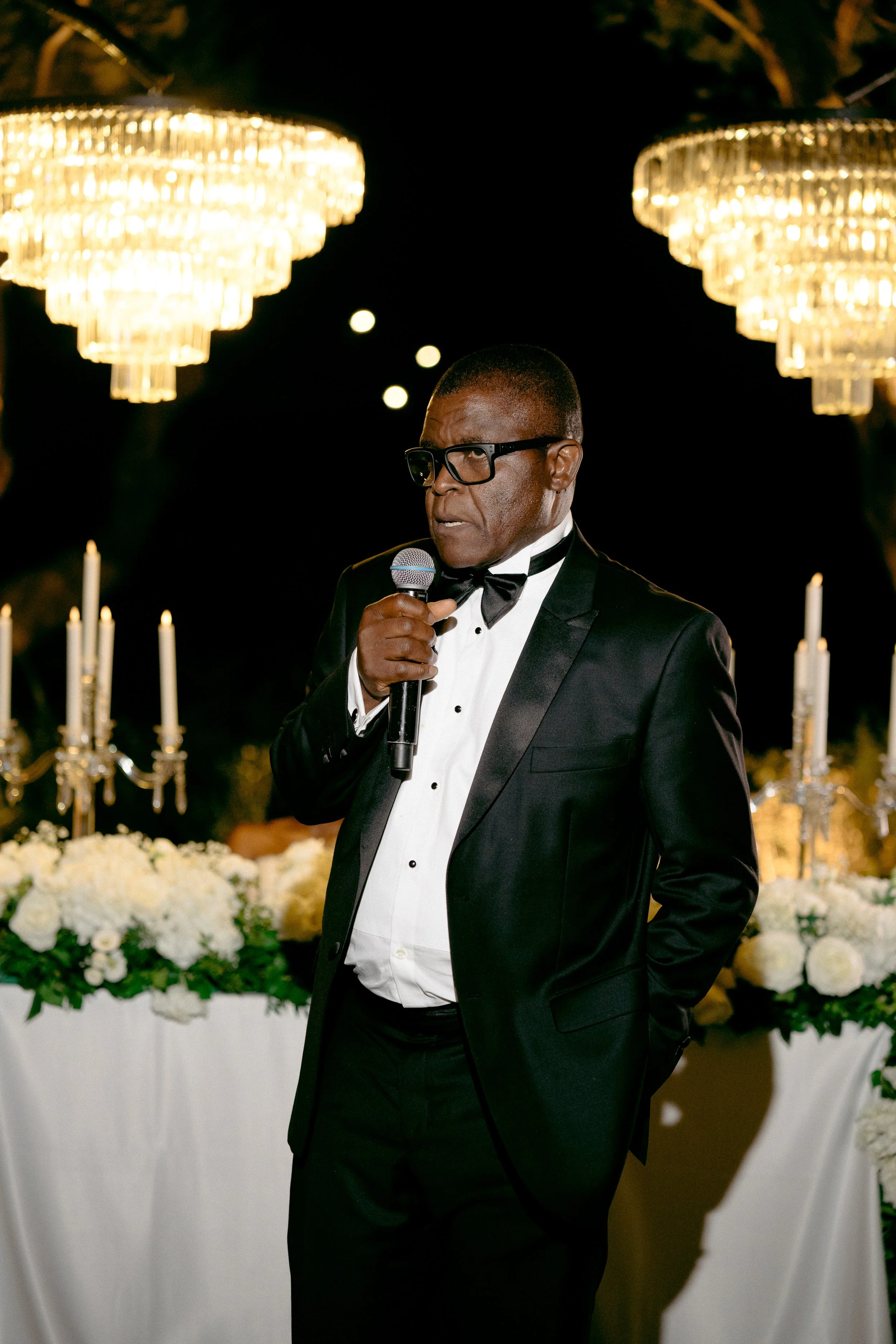 A man in a tuxedo and glasses holding a microphone at an elegant evening event with chandeliers, candles, and white flowers.