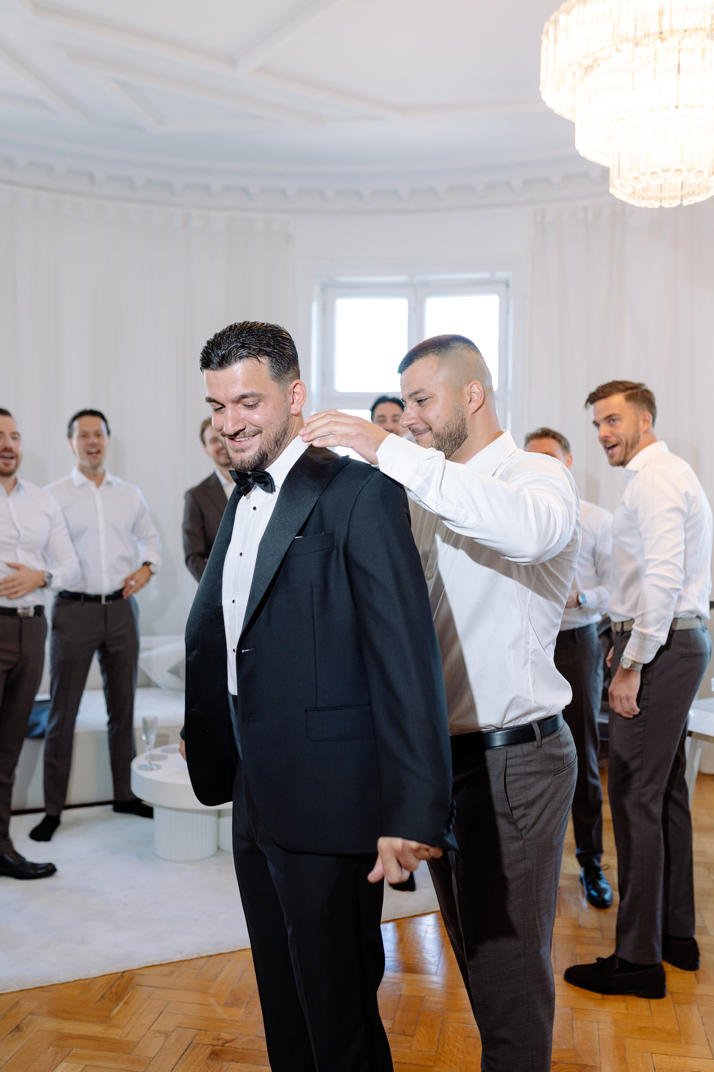 Groom in black tuxedo with bowtie getting dressed by a man behind him at a wedding or formal event, with other groomsmen in white shirts and dark trousers in the background.