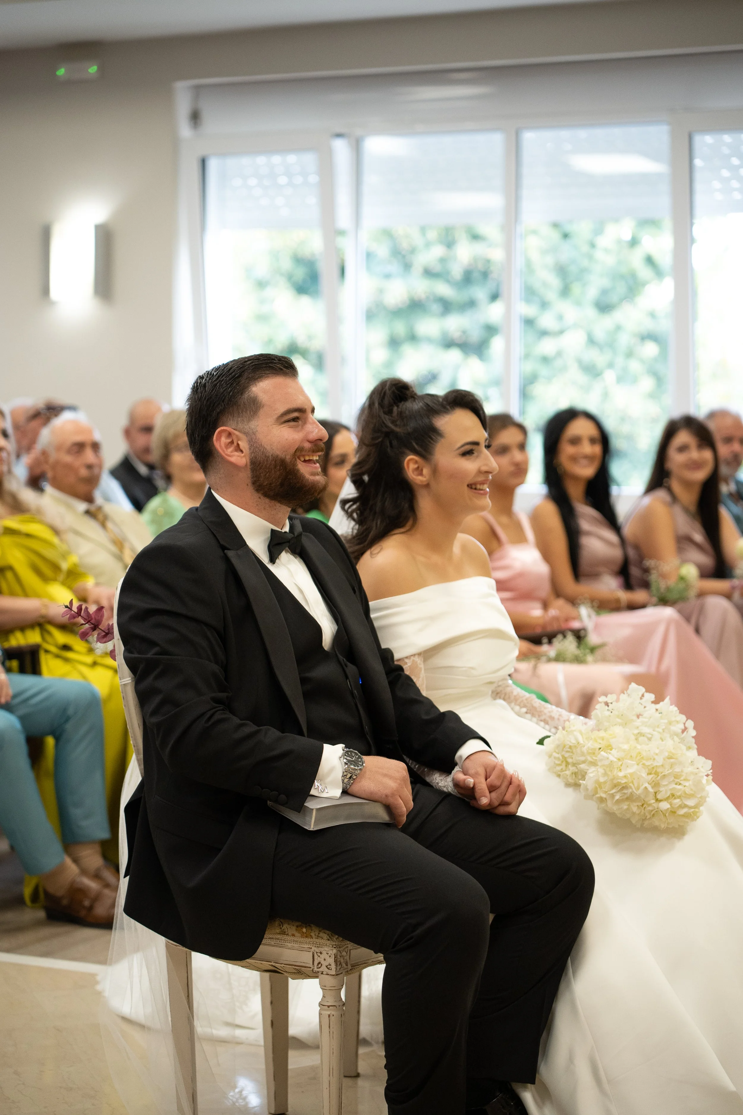 A bride and groom sitting together during their wedding ceremony, smiling happily, with guests seated behind them.