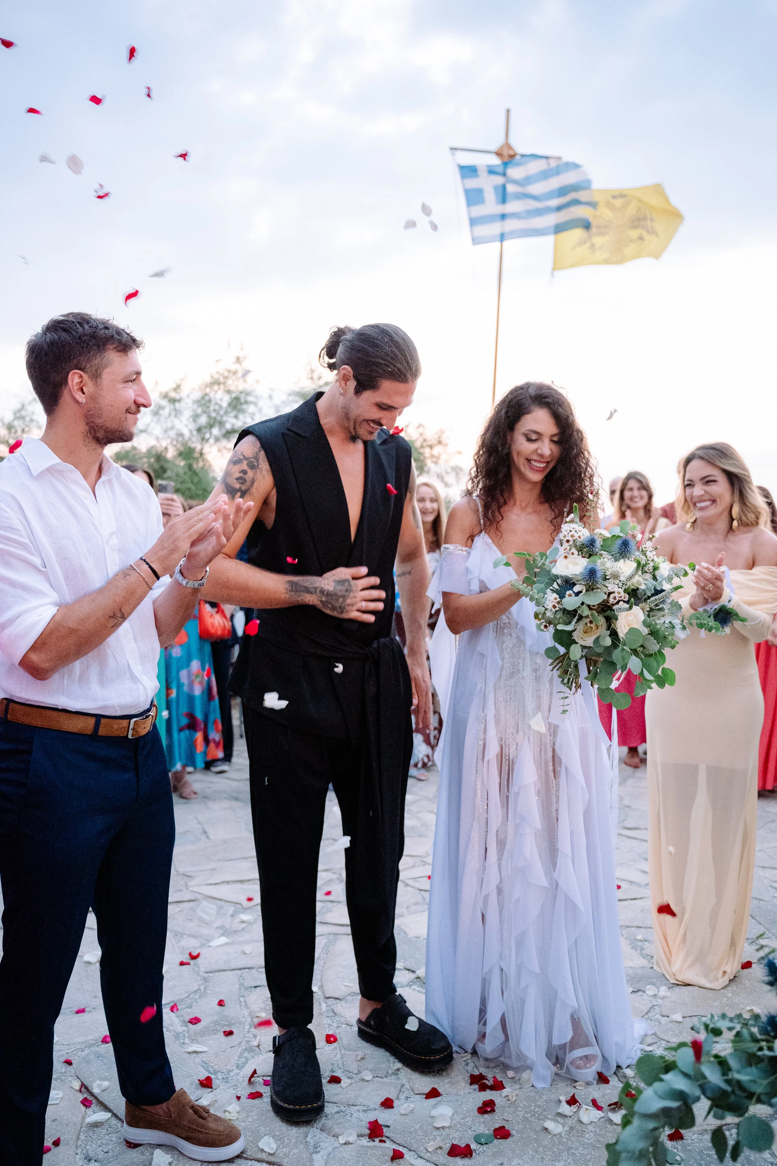 Couple getting married with friends celebrating outdoors, holding a bouquet, with Greek and Greek Orthodox flags in the background, and flower petals on the ground.