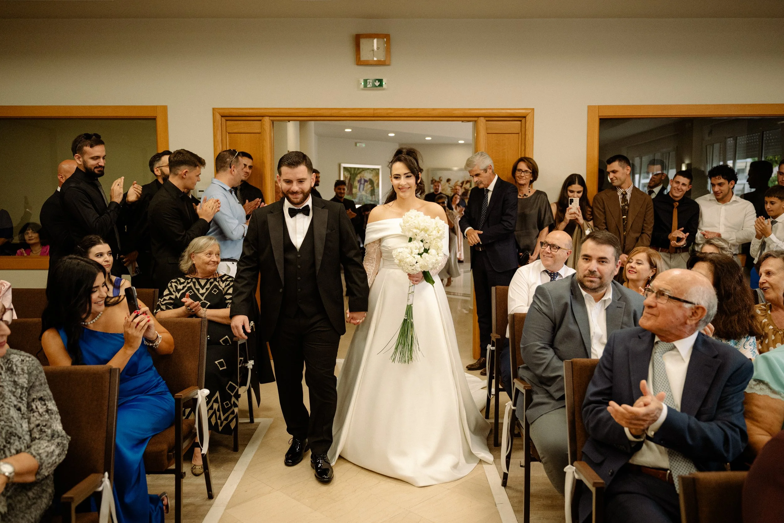 A bride and groom walking down the aisle, holding hands, as guests applaud and take pictures in a wedding ceremony.