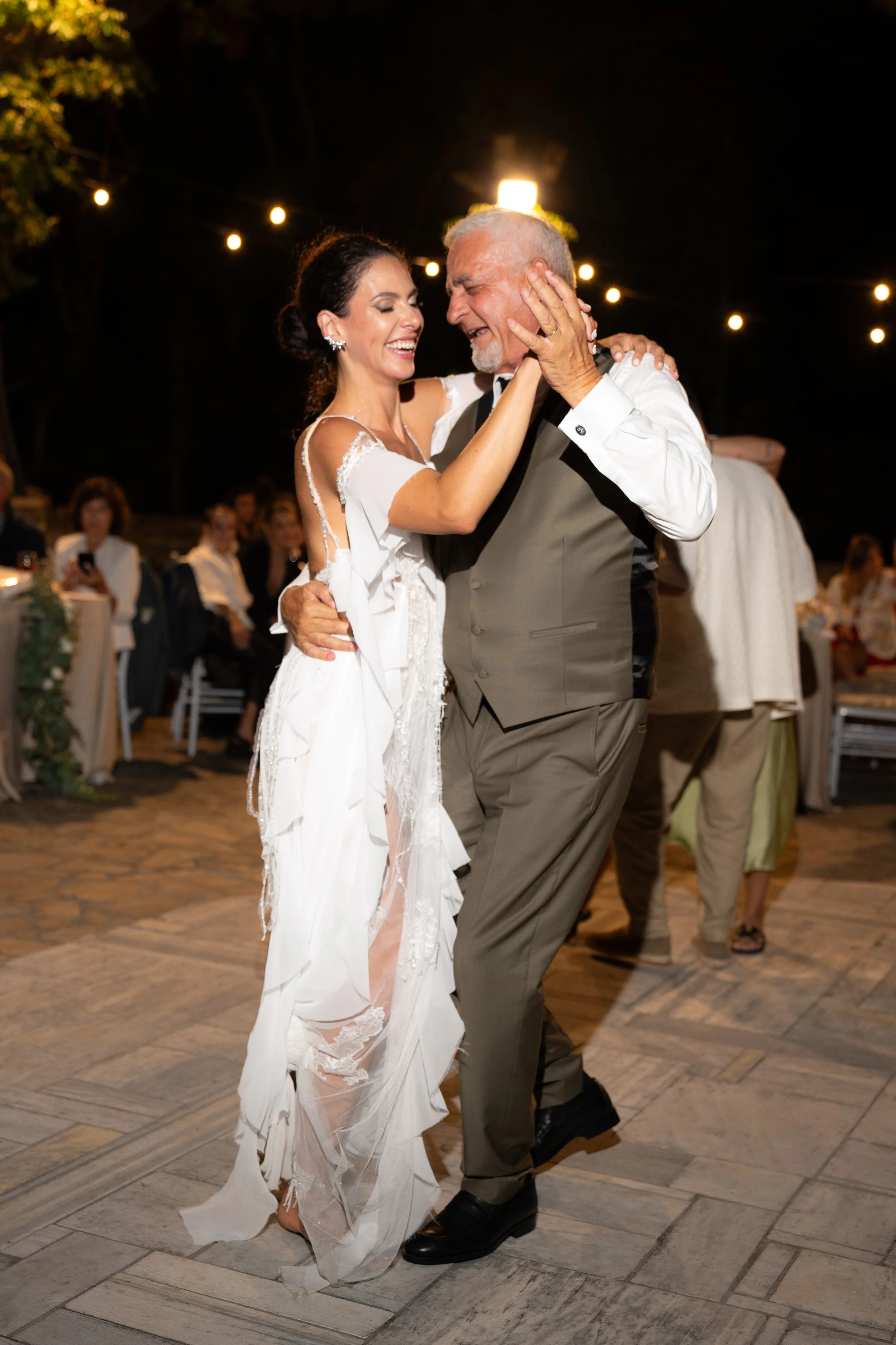 A bride and an older man, possibly her father, dancing happily at a wedding reception outdoors at night, under string lights.