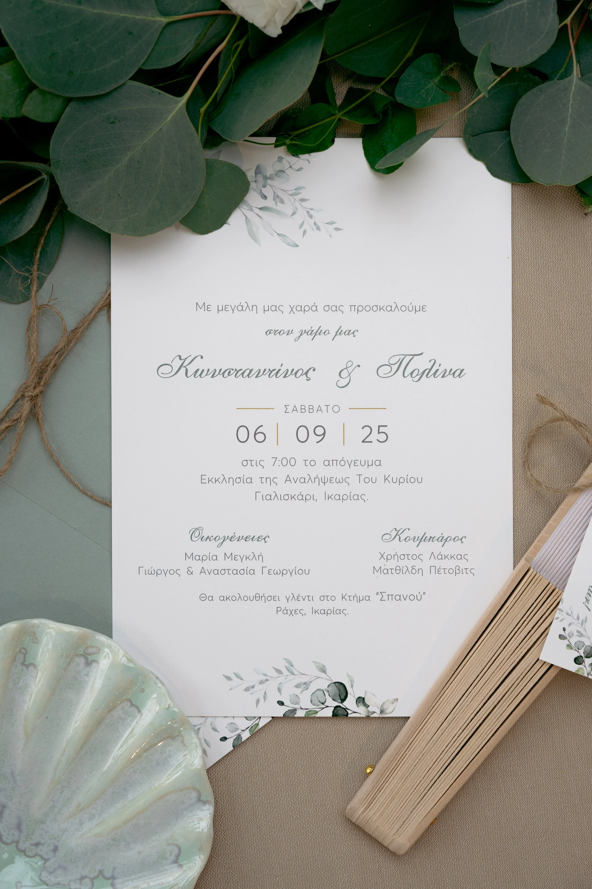 A wedding invitation card decorated with green leaves, placed on a beige surface with a ceramic dish and a wooden fan nearby.