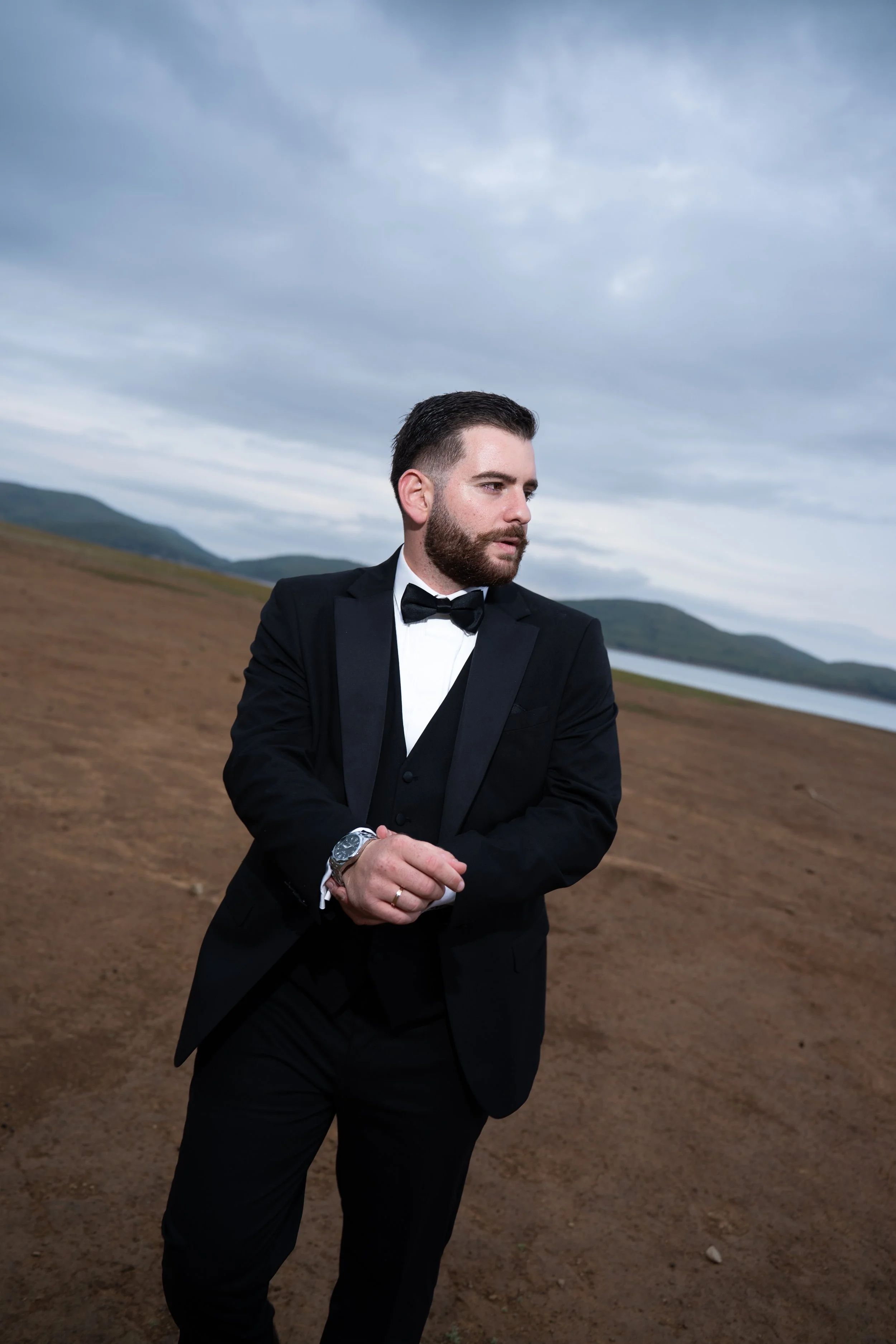 A man in a black tuxedo and bow tie stands outdoors on a dirt path near water and mountains, looking to the side with a serious expression.