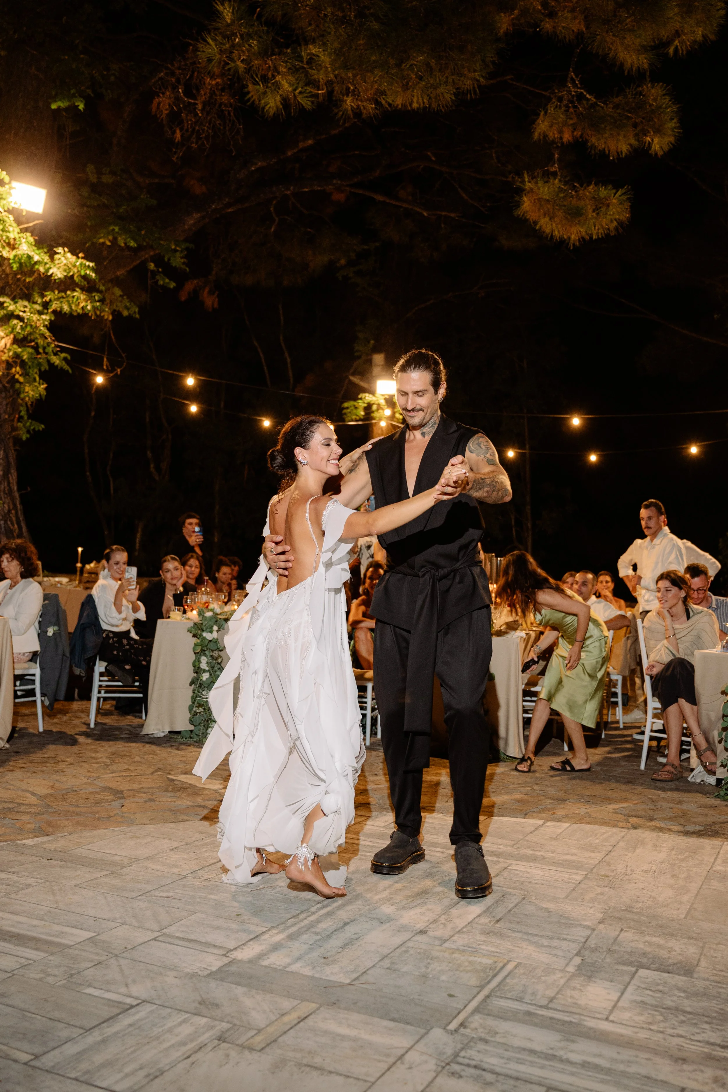 A bride and groom are dancing together at their outdoor wedding reception at night, surrounded by seated guests, string lights, and trees.