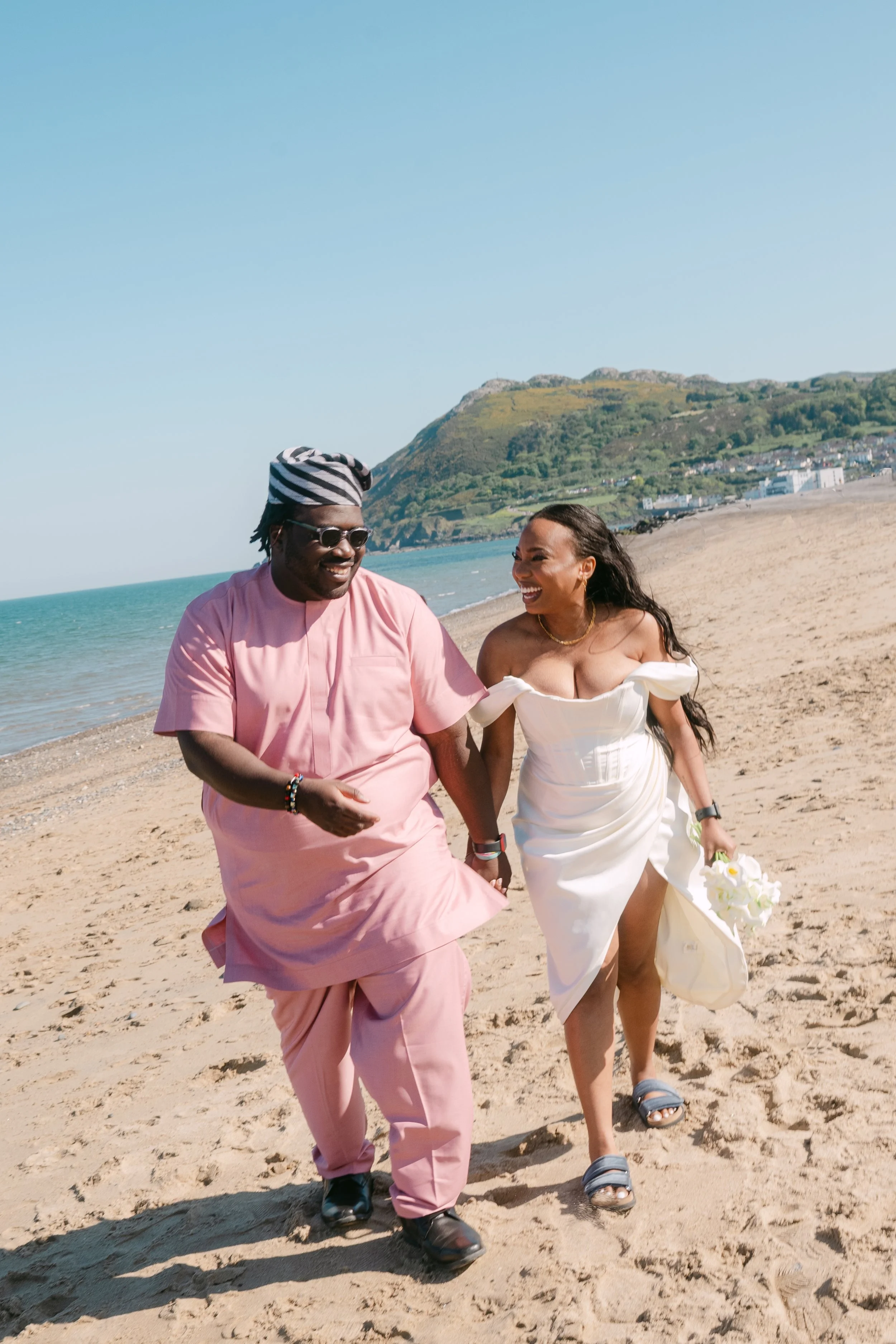 A couple walking on the beach, smiling and holding hands, with a scenic coastline and green hills in the background.