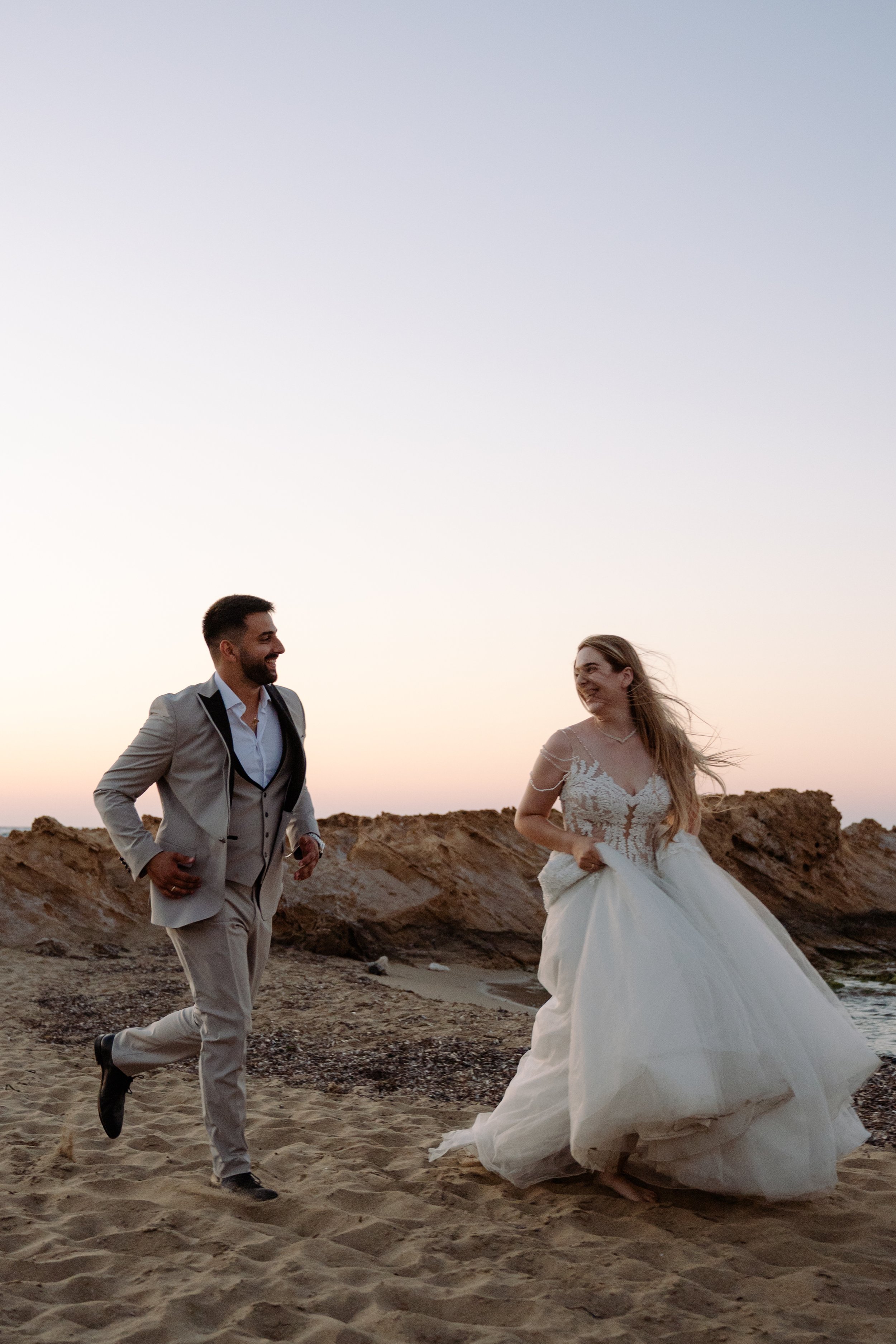 A newlywed couple running on a beach at sunset, with rocky formations in the background. The groom is in a light gray suit, and the bride is in a white wedding gown.