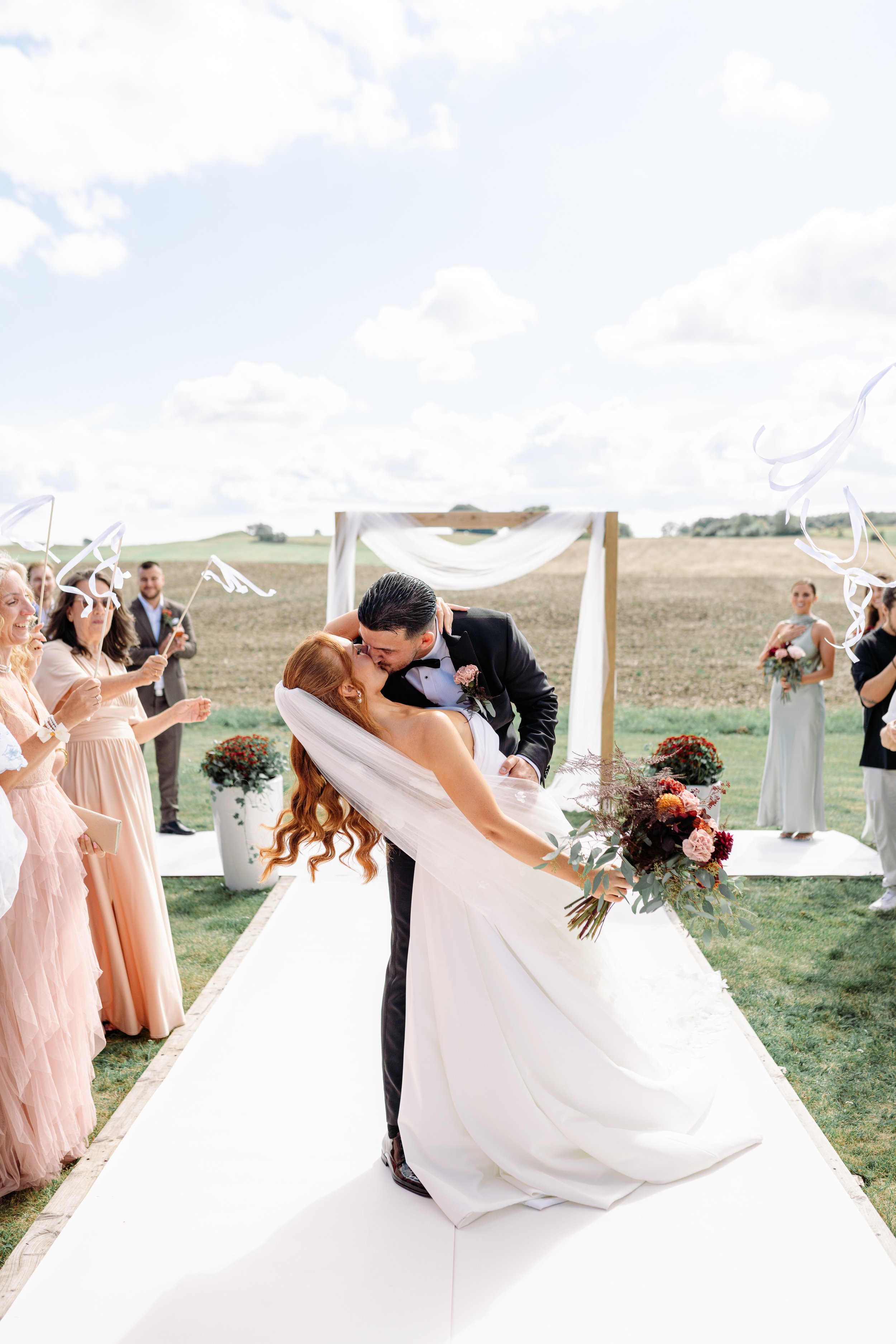 A newlywed couple sharing a kiss at an outdoor wedding ceremony, with friends and family celebrating and celebrating around them.