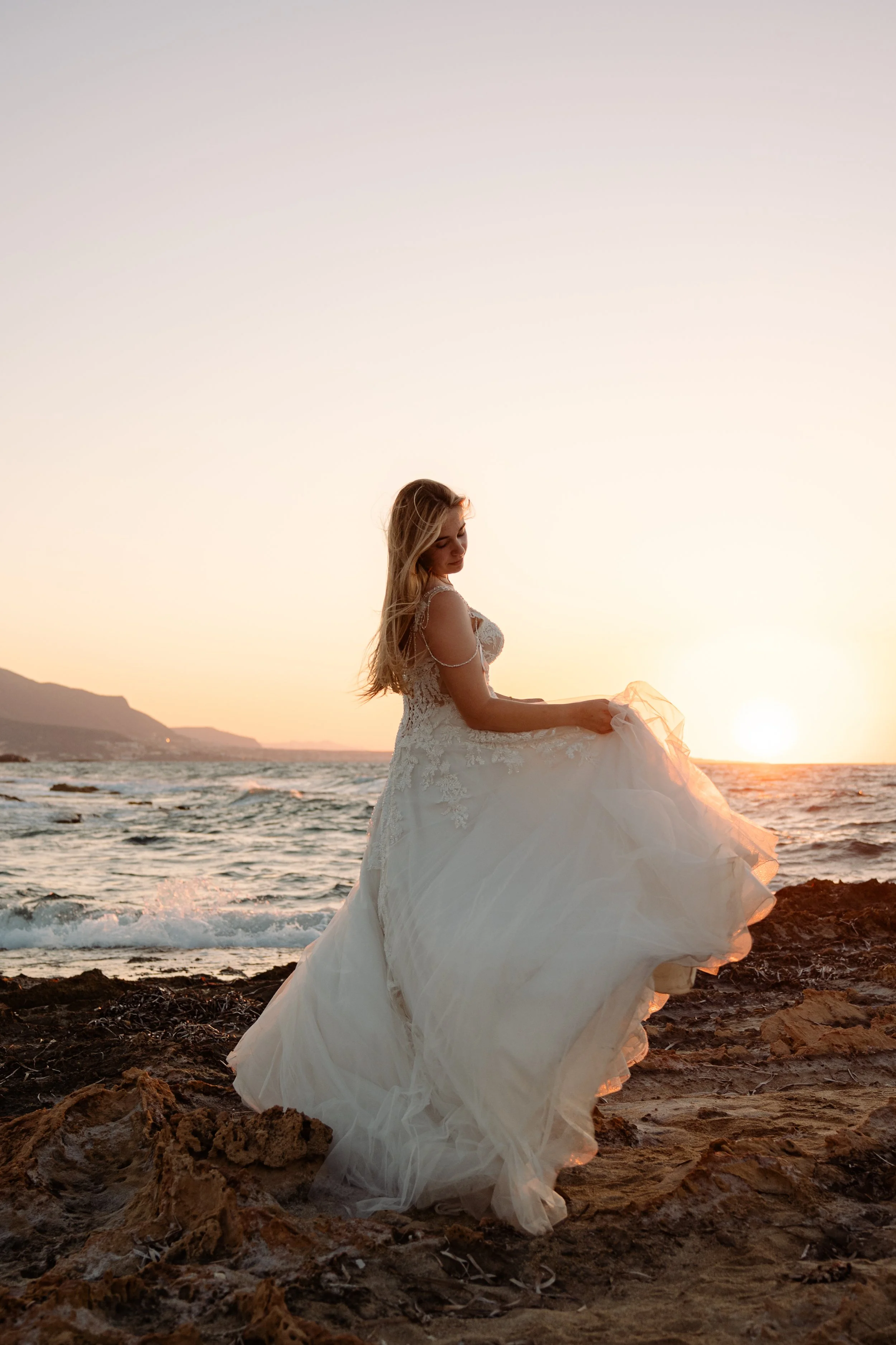 A bride in a white wedding gown standing on a rocky beach at sunset.
