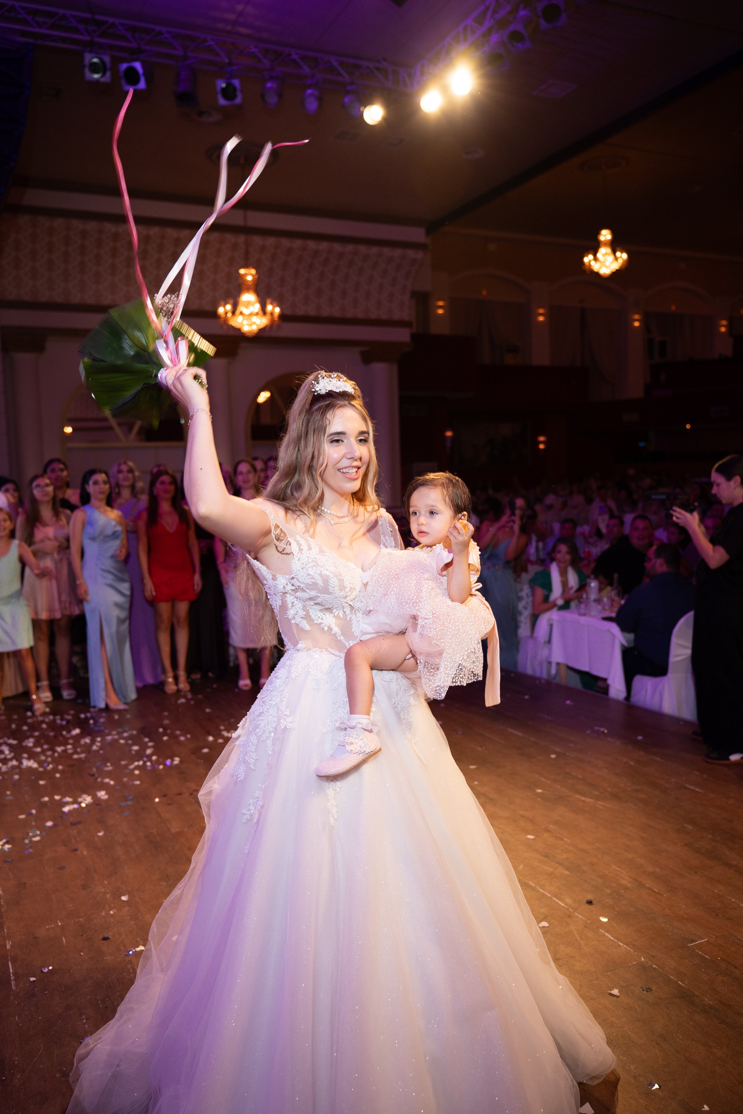 A bride in a white wedding gown holding a bouquet and a young girl in a pink dress at a wedding reception with guests, chandeliers, and a dance floor.