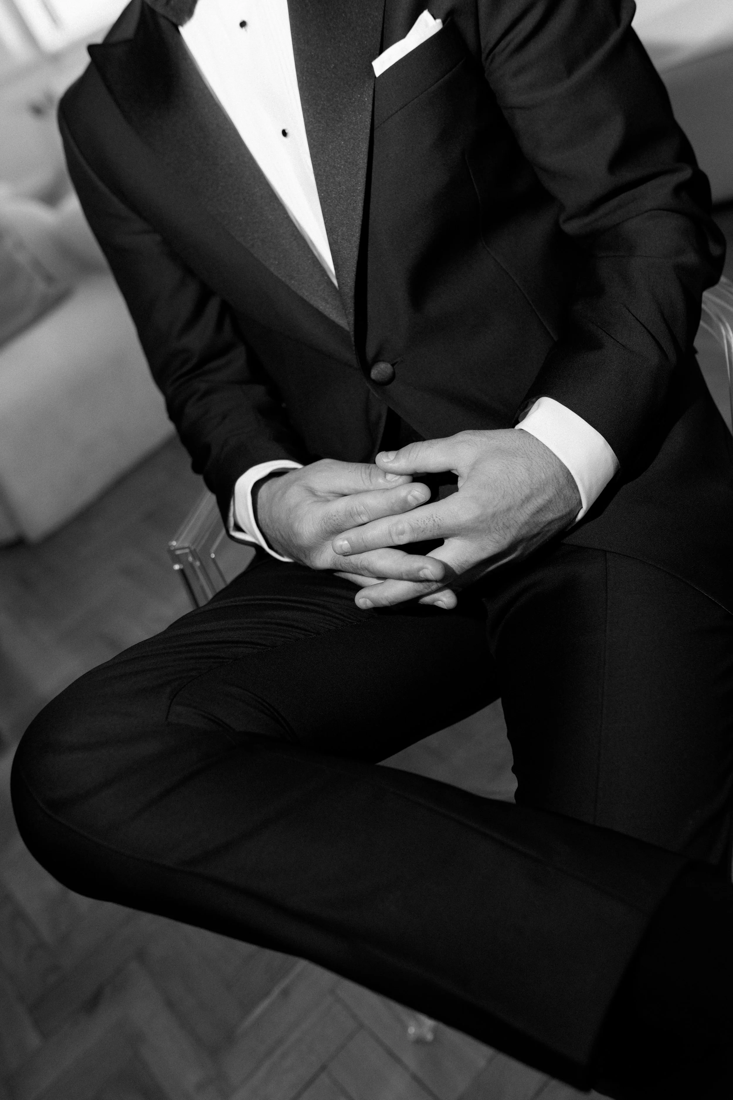Black and white photo of a man in a tuxedo with a bow tie, sitting with his hands clasped on his lap.